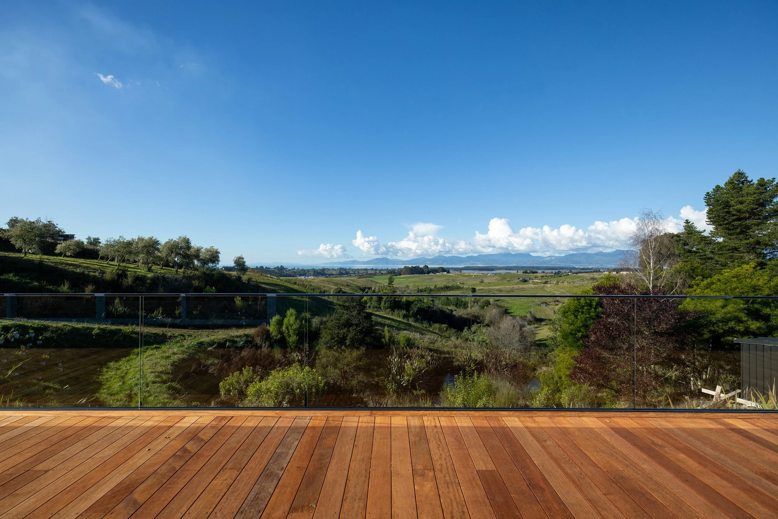 A view from a wooden deck overlooking a green landscape with rolling hills, trees, and mountains in the distance under a blue sky with some clouds.