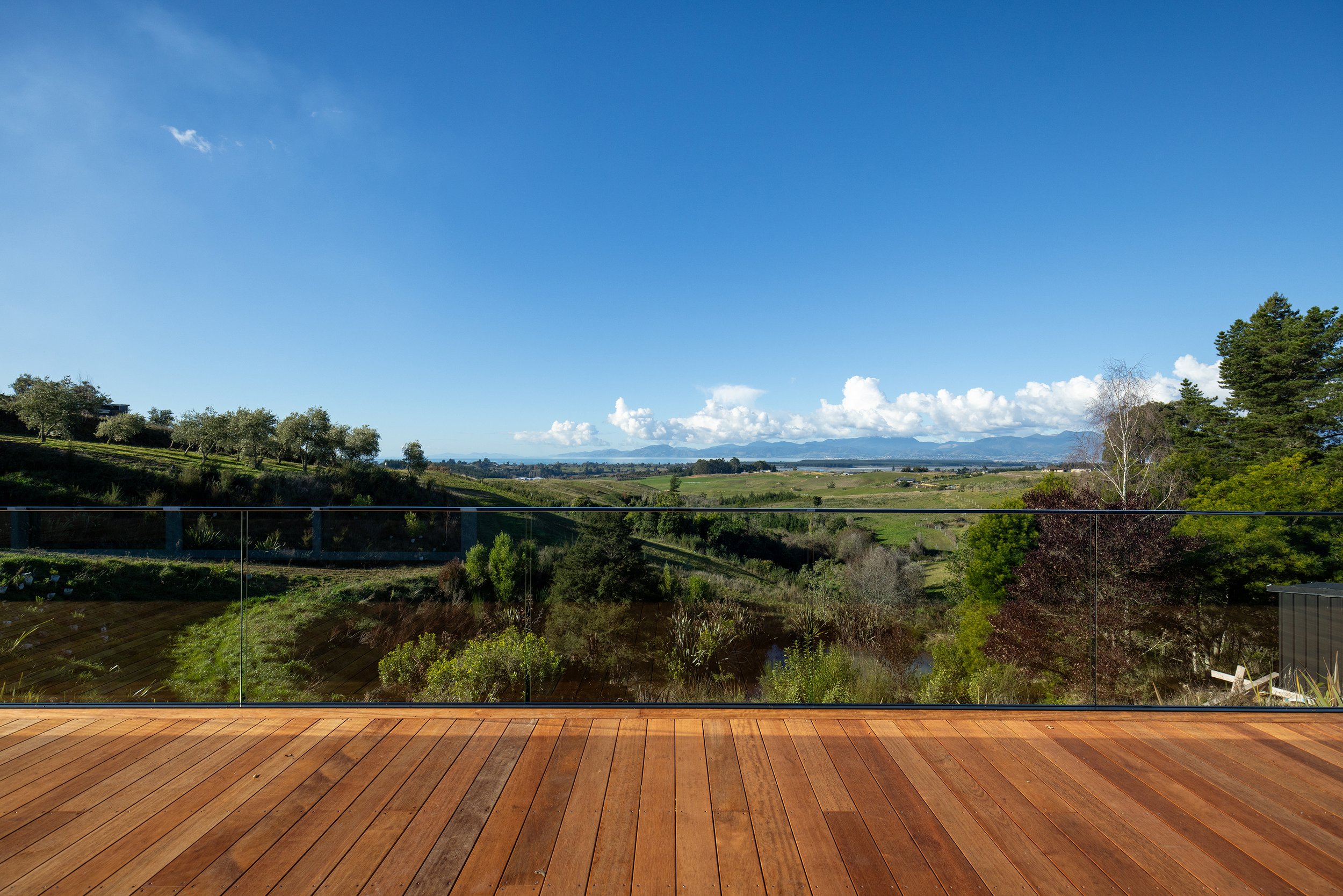 A view from a wooden deck overlooking a green landscape with rolling hills, trees, and mountains in the distance under a blue sky with some clouds.