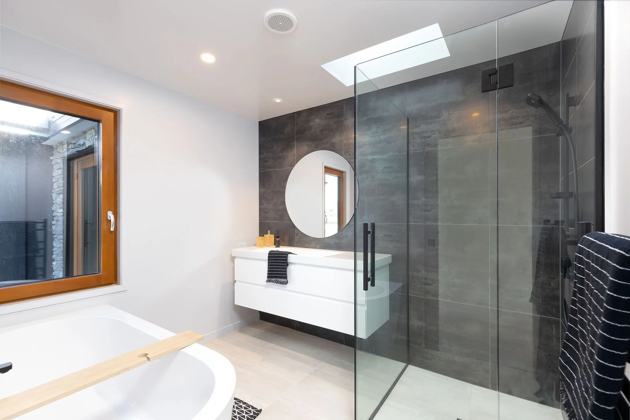 Modern bathroom with a white bathtub, a black and white striped towel, a black shower area with glass doors, a white vanity with round mirror, and a window with wood trim.