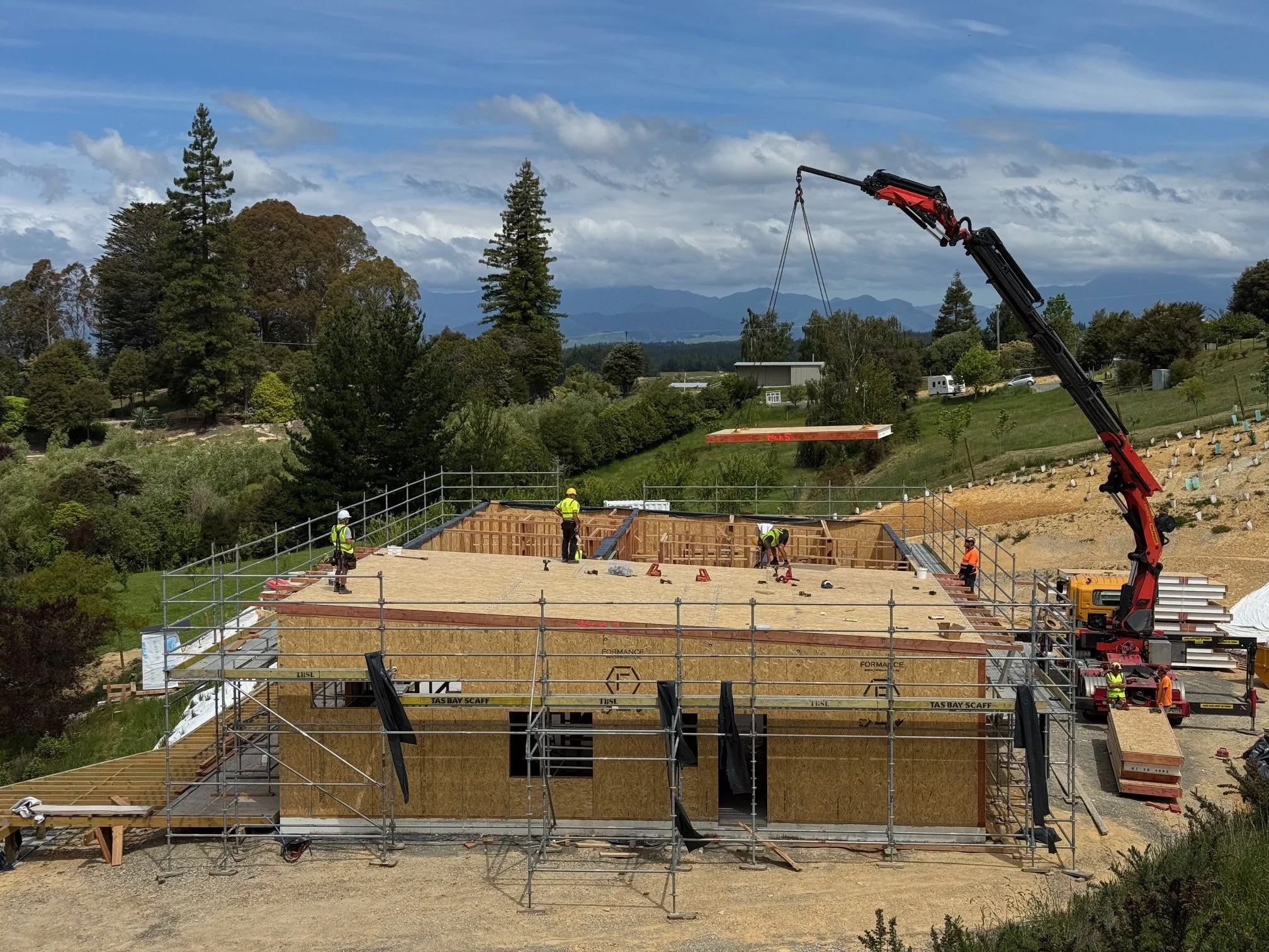 Construction workers building a house with wooden framework and scaffolding, and a crane lifting a large wooden panel at a rural construction site.