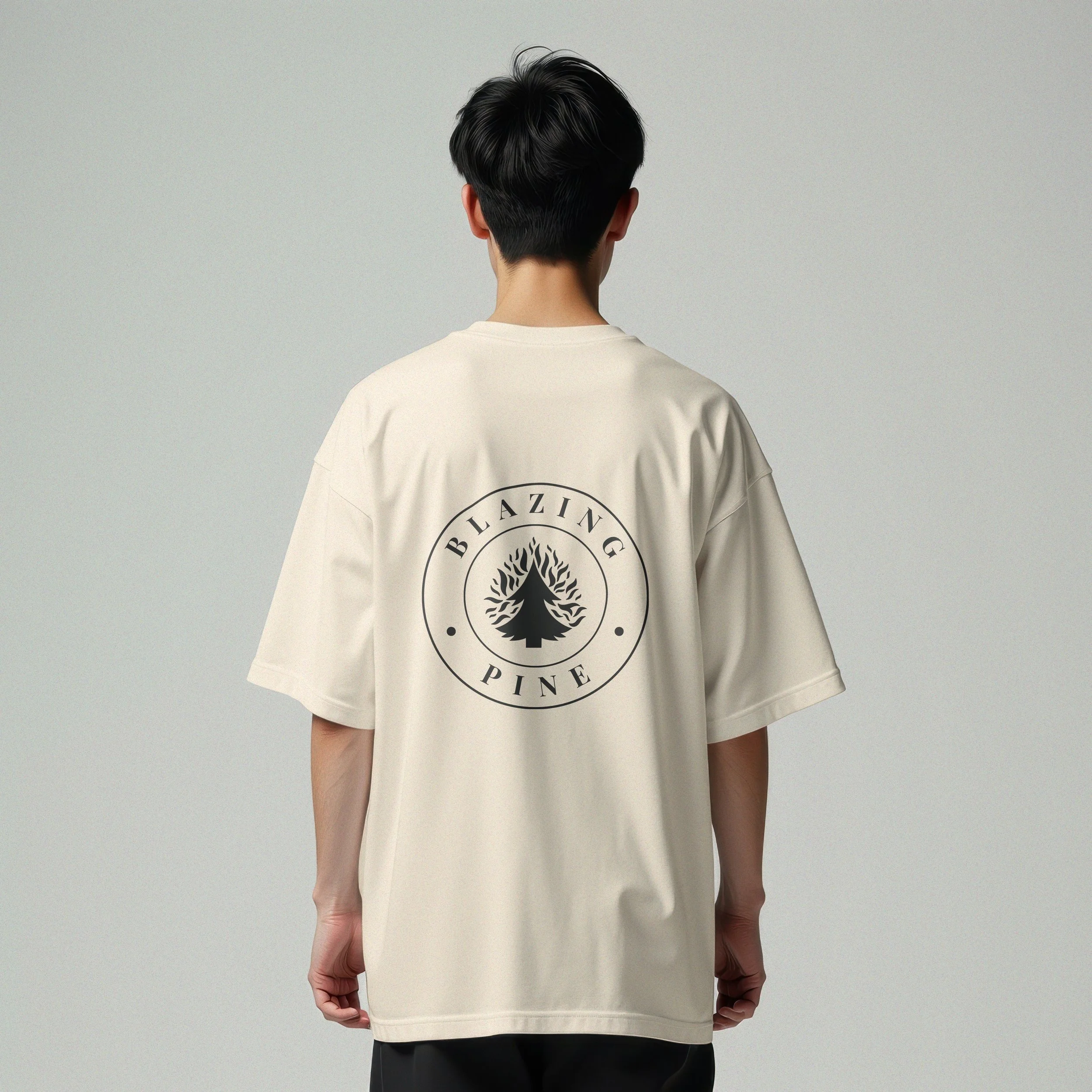 Back of a person wearing a beige oversized t-shirt with a black circular logo featuring a burning pine tree, with the words "BLAZING PINE" around it.