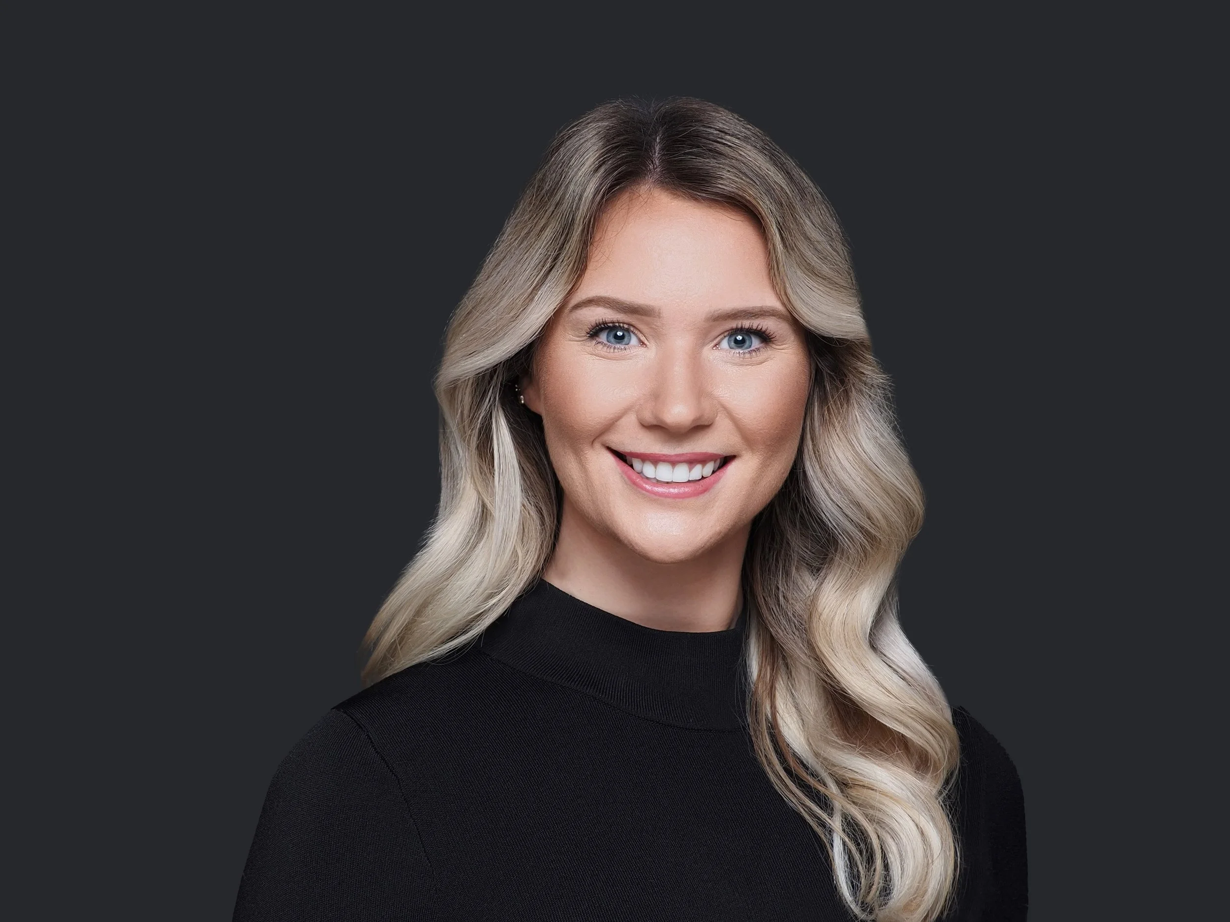 A portrait of a smiling woman with long, wavy blonde hair, blue eyes, and fair skin, wearing a black top, against a dark background.