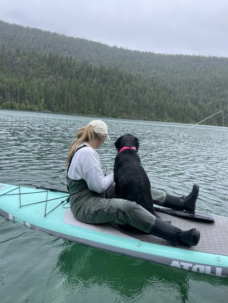 A woman in outdoor clothing and a cap sitting on a paddleboard with a black dog, fishing in a lake surrounded by forested hills.