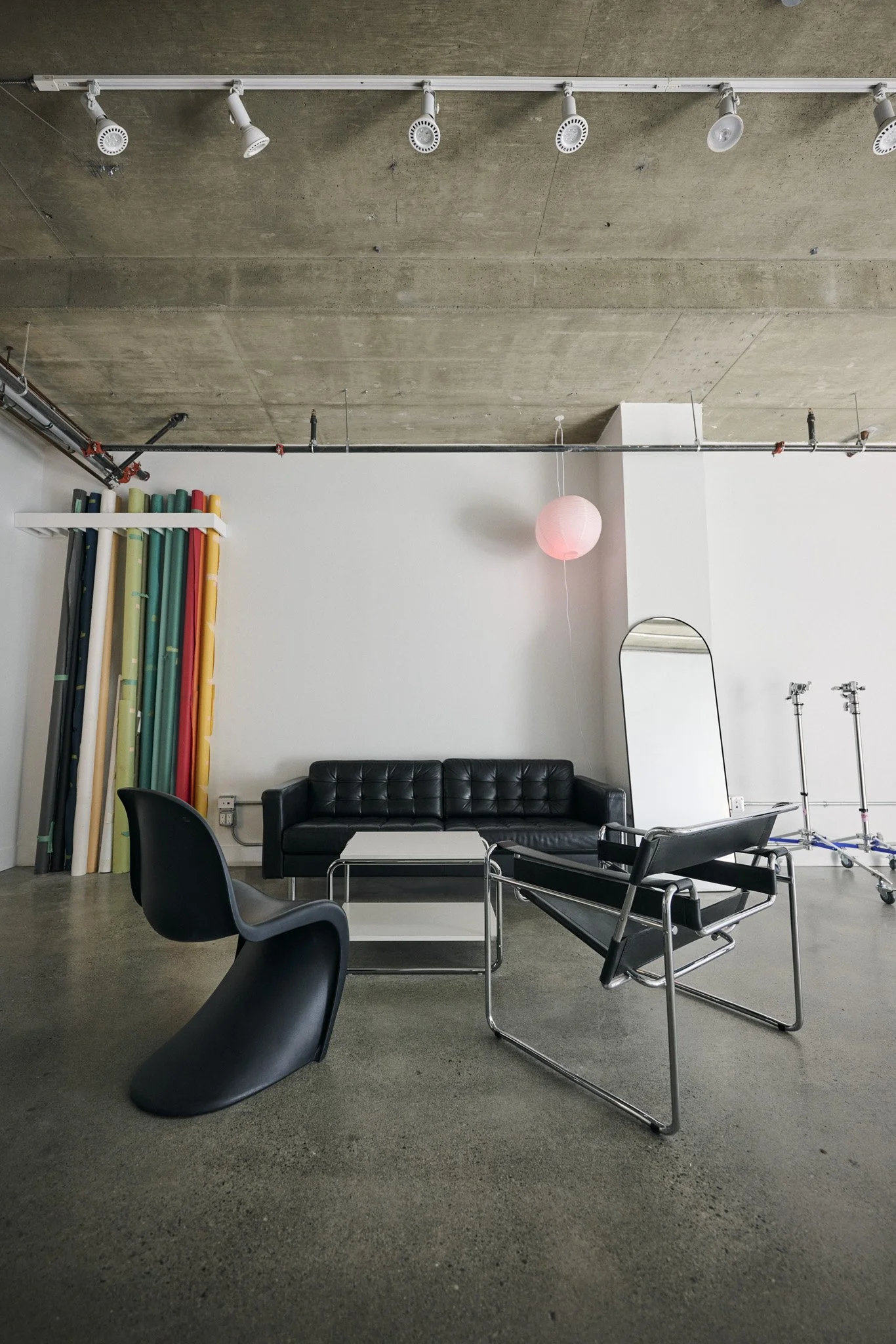 Interior office or studio space with a concrete ceiling, track lighting, and various furniture including a black leather sofa, black chairs, a white coffee table, and a black lounge chair. There are tall colorful poles on the left, a pink paper lantern hanging from the ceiling, a mirror on the right, and some pipes and equipment.