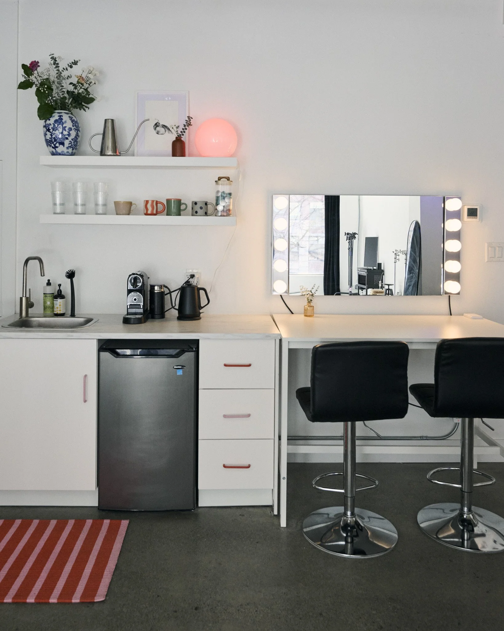 Modern kitchen with white countertops, black and white coffee machines, a small fridge, two bar stools, a large mirror with vanity lights, and shelves with decorative items and dishes.