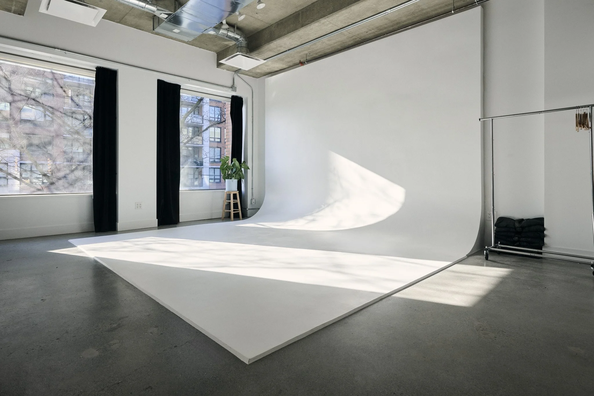 Empty photography studio with large windows, a white backdrop, a potted plant on a stool, black curtains, and minimalist decor.