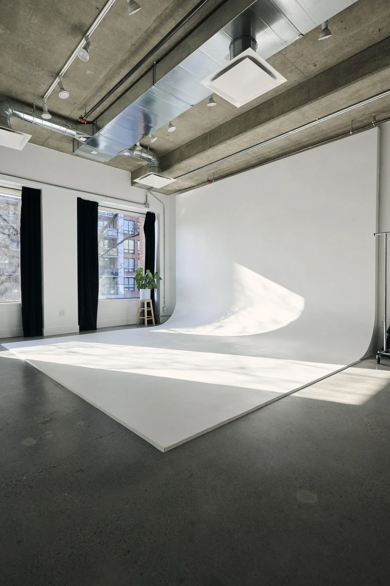 Empty photography studio with white backdrop, black curtains, window showing buildings outside, a potted plant, and ceiling vents.