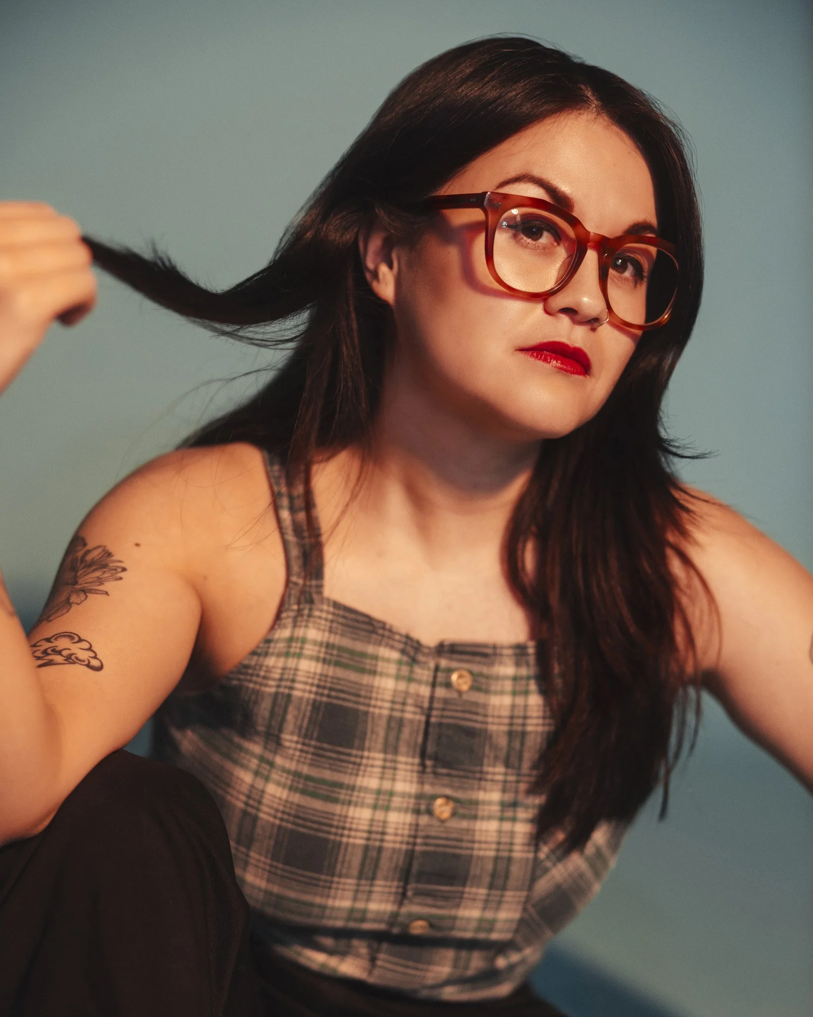 Close-up of a woman with dark hair, wearing tortoiseshell glasses and red lipstick, holding a strand of her hair.