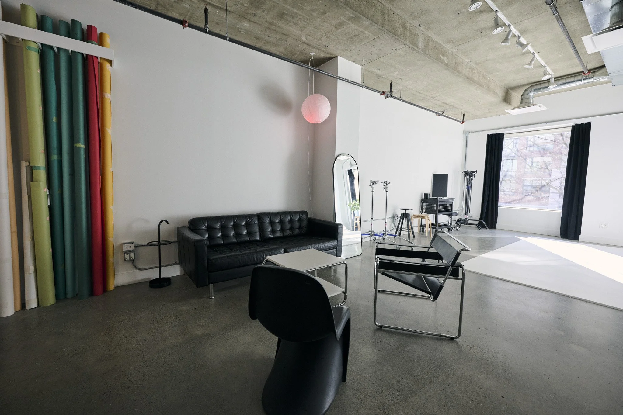 A spacious room with an industrial style ceiling featuring exposed concrete and ductwork. The room contains a black leather sofa, a small white table, black chairs, and a full-length mirror. There's a large window with black curtains allowing natural light to fill the space, and equipment such as light stands and photography gear is visible, indicating it may be a studio or creative workspace.