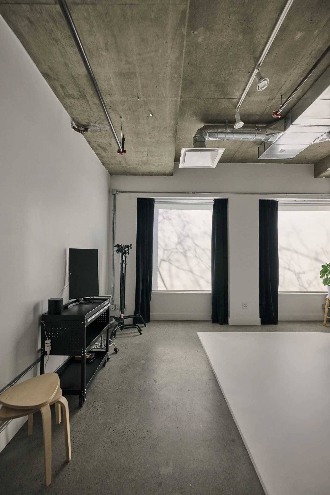 An industrial-style room with a concrete ceiling, black curtains over a large window, and modern ventilation ducts. To the left, there is a small black TV on a black rolling cart, with equipment below. Next to the TV, two wooden stools are placed. A white backdrop is set up on the right side, possibly for photo shoots or filming.