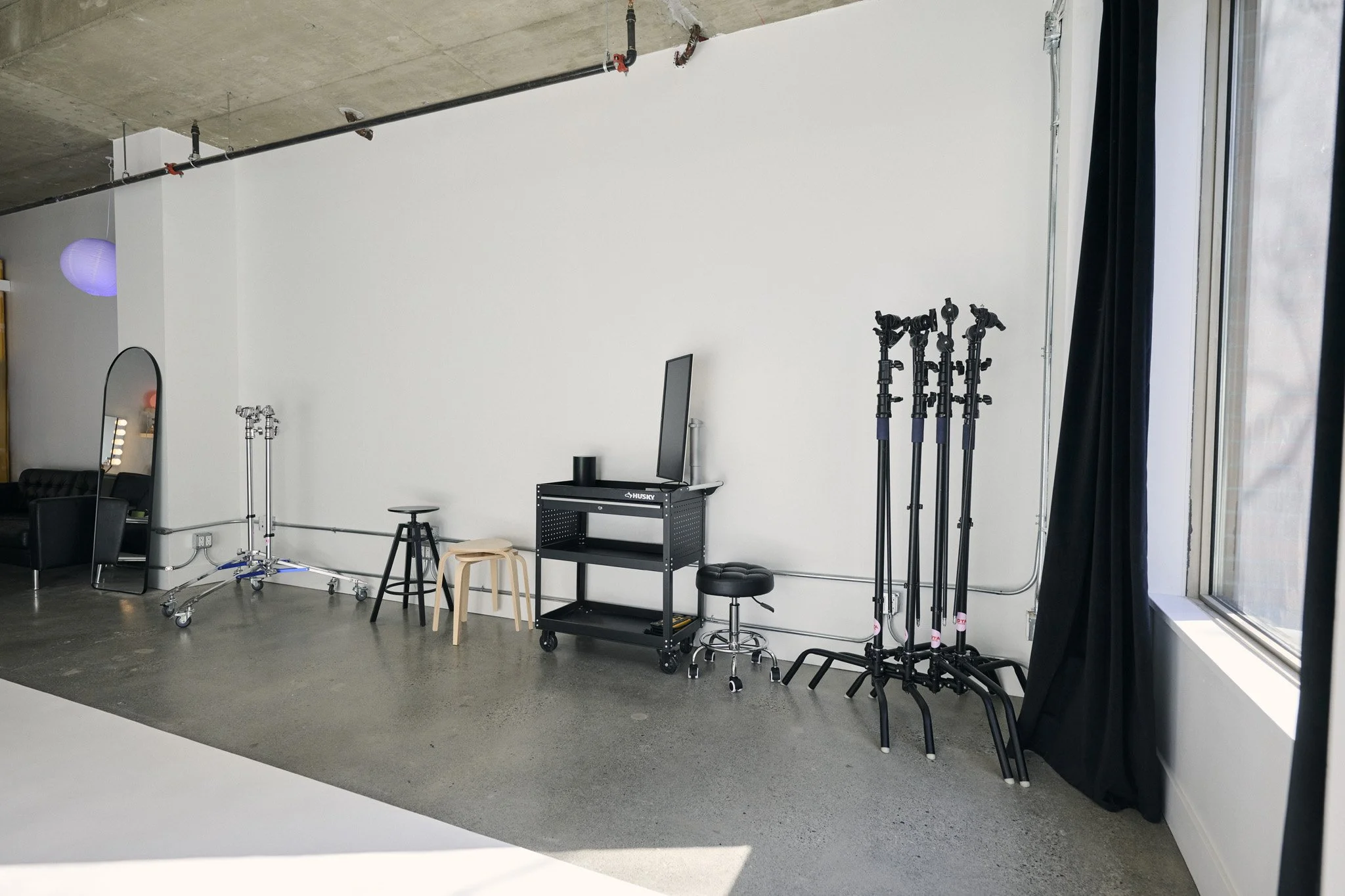 Photography studio with equipment including light stands, a mirror, chairs, a rolling cart, and a black curtain by a large window with a city view.