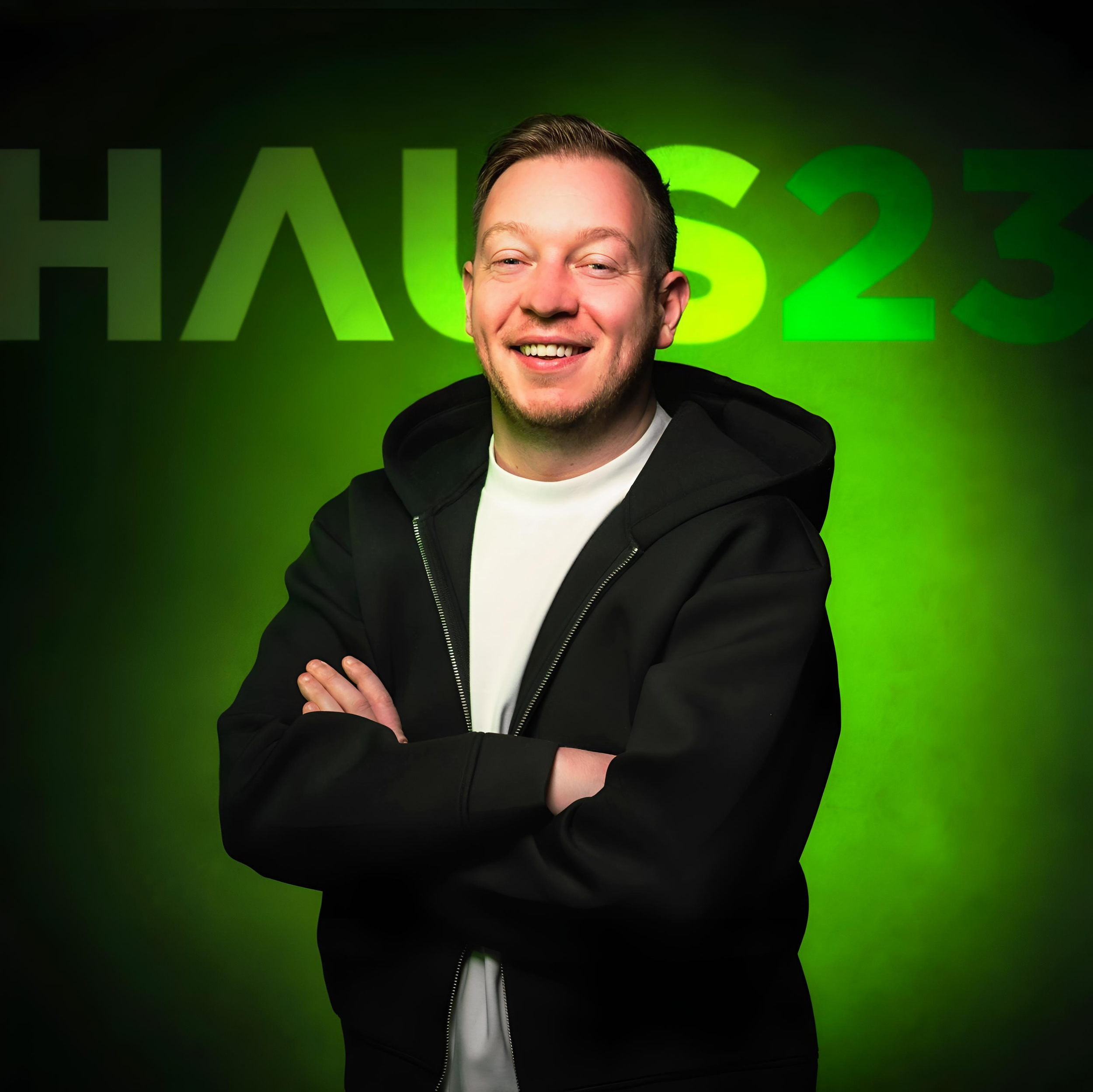 A smiling man with folded arms standing in front of a green background with the words 'HALLS 23' on it.