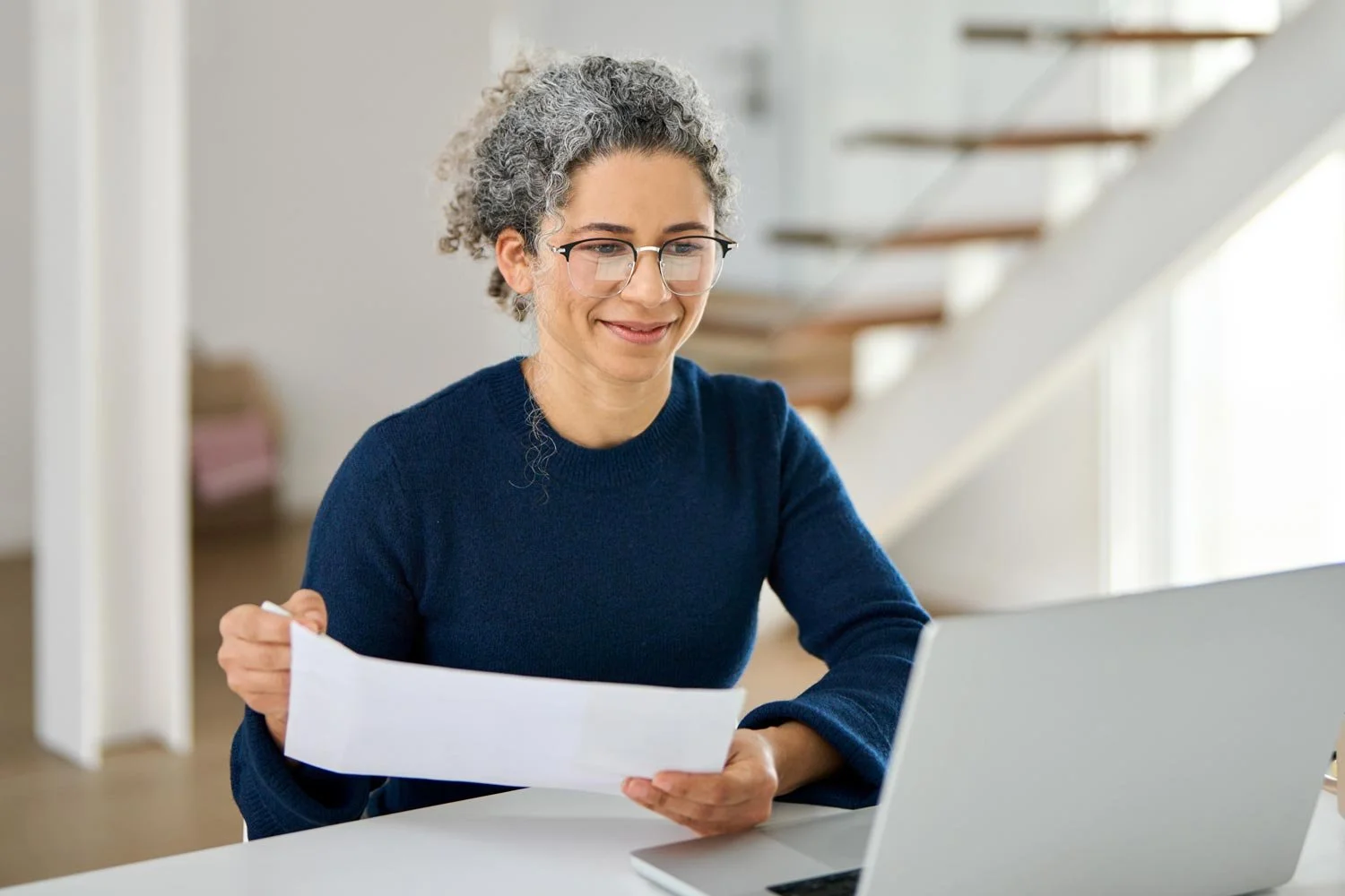 Smiling mature woman in dark blue shirt holding a document looks toward a laptop screen on the desk.