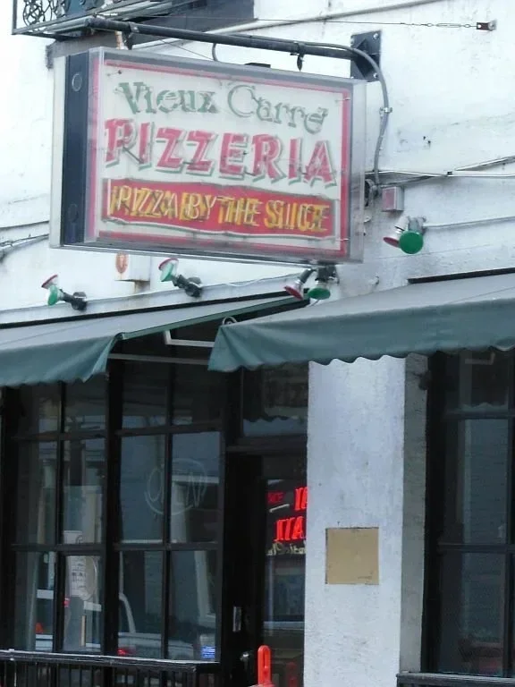 Exterior of a pizzeria with a sign reading "Vieux Carré Pizzeria" and "Pizza by the Slice". The building has storefront windows and green awnings.