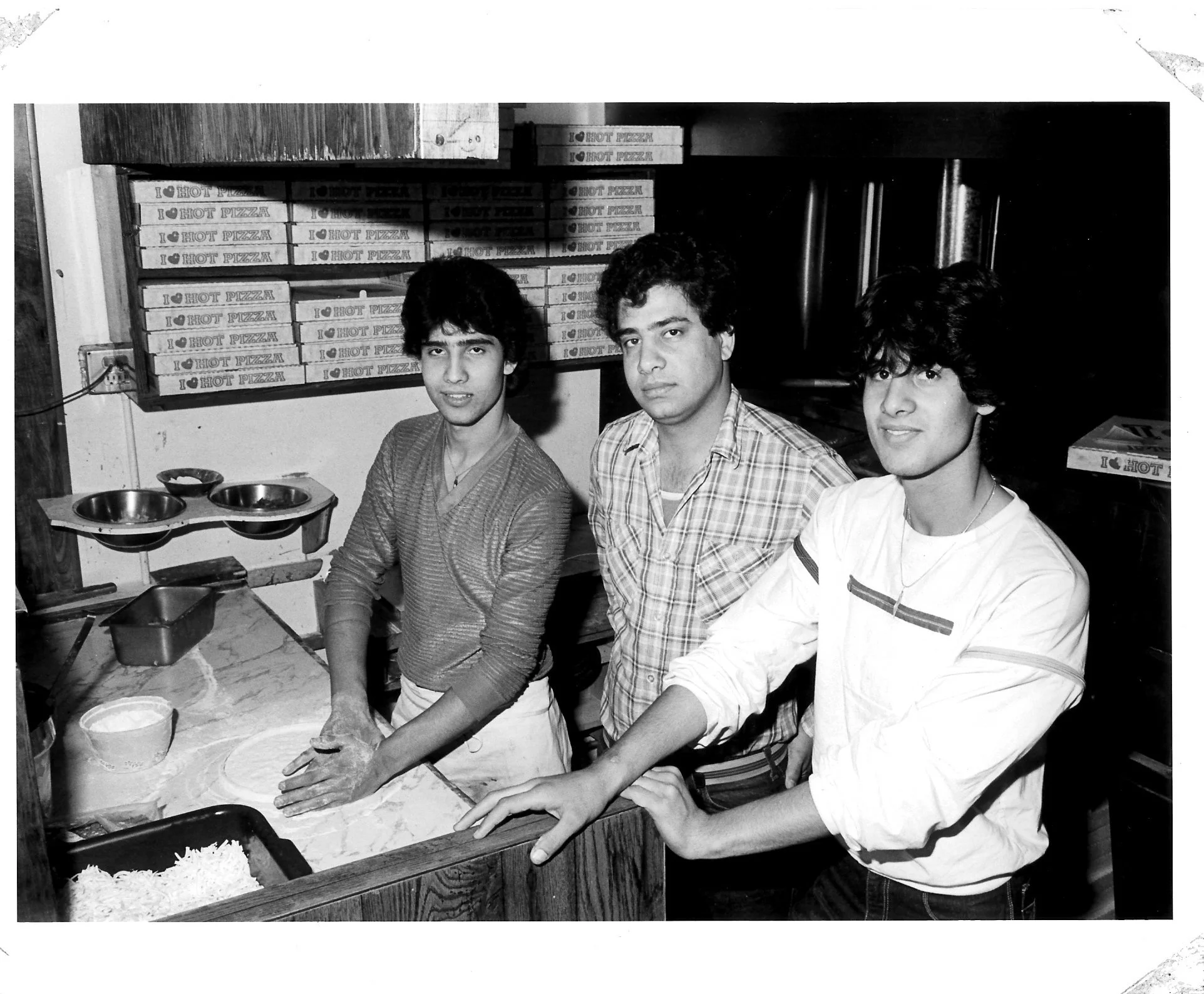 Three young men with dark hair behind a counter in a pizza restaurant, with stacked pizza boxes labeled "I ❤️ HOT PIZZA" on the shelves behind them.