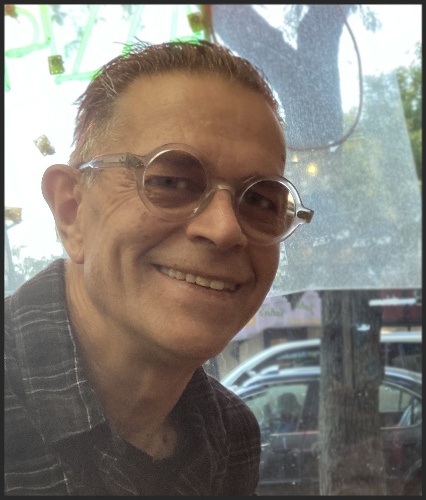 A smiling Chris Cloutier with short, slicked-back hair and round tinted glasses, wearing a dark plaid shirt, sits indoors near a window with trees and parked cars visible outside.