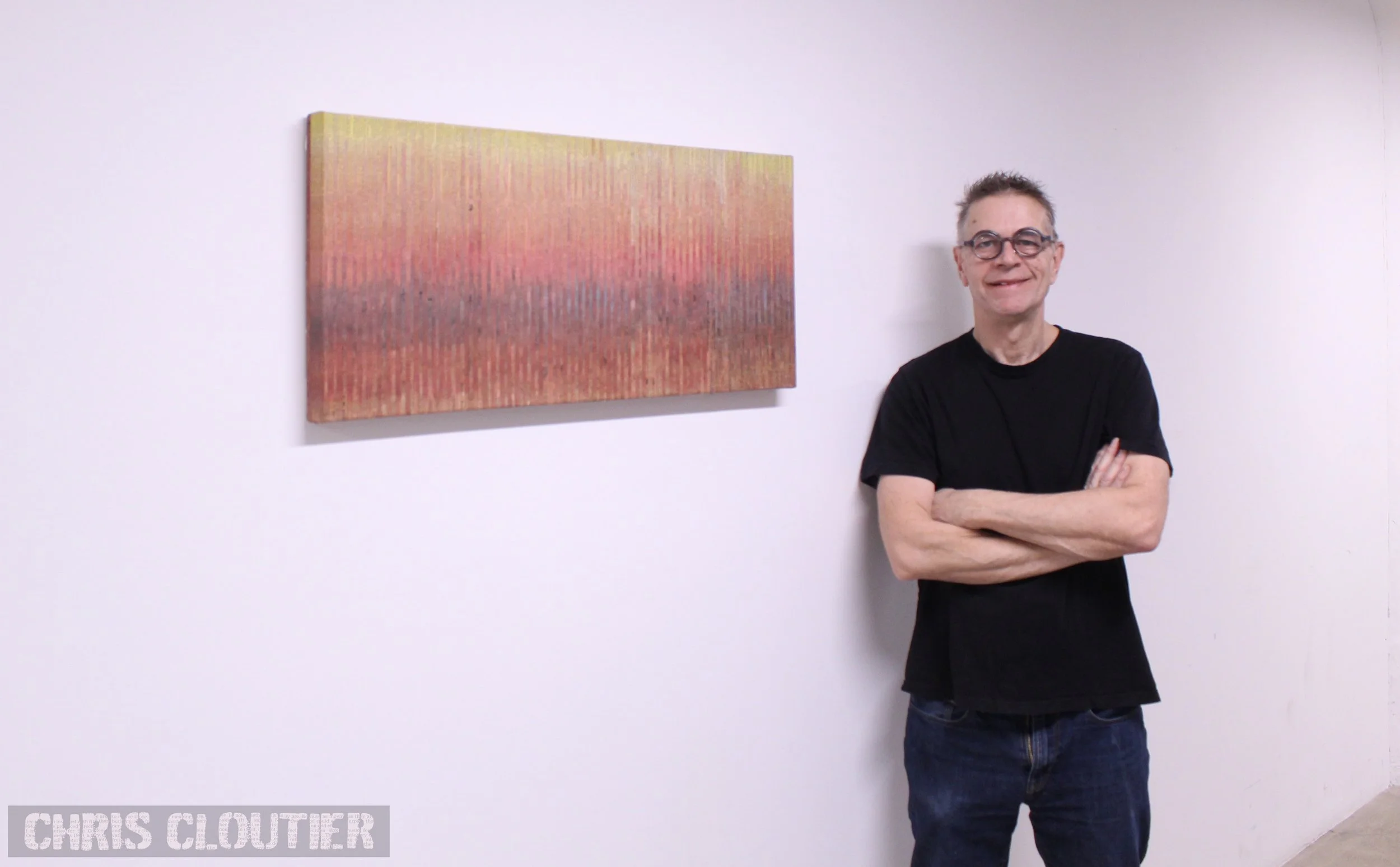 Chris Cloutier stands with glasses, wearing a black t-shirt and jeans, standing with arms crossed next to a colorful abstract collage on a white gallery wall.