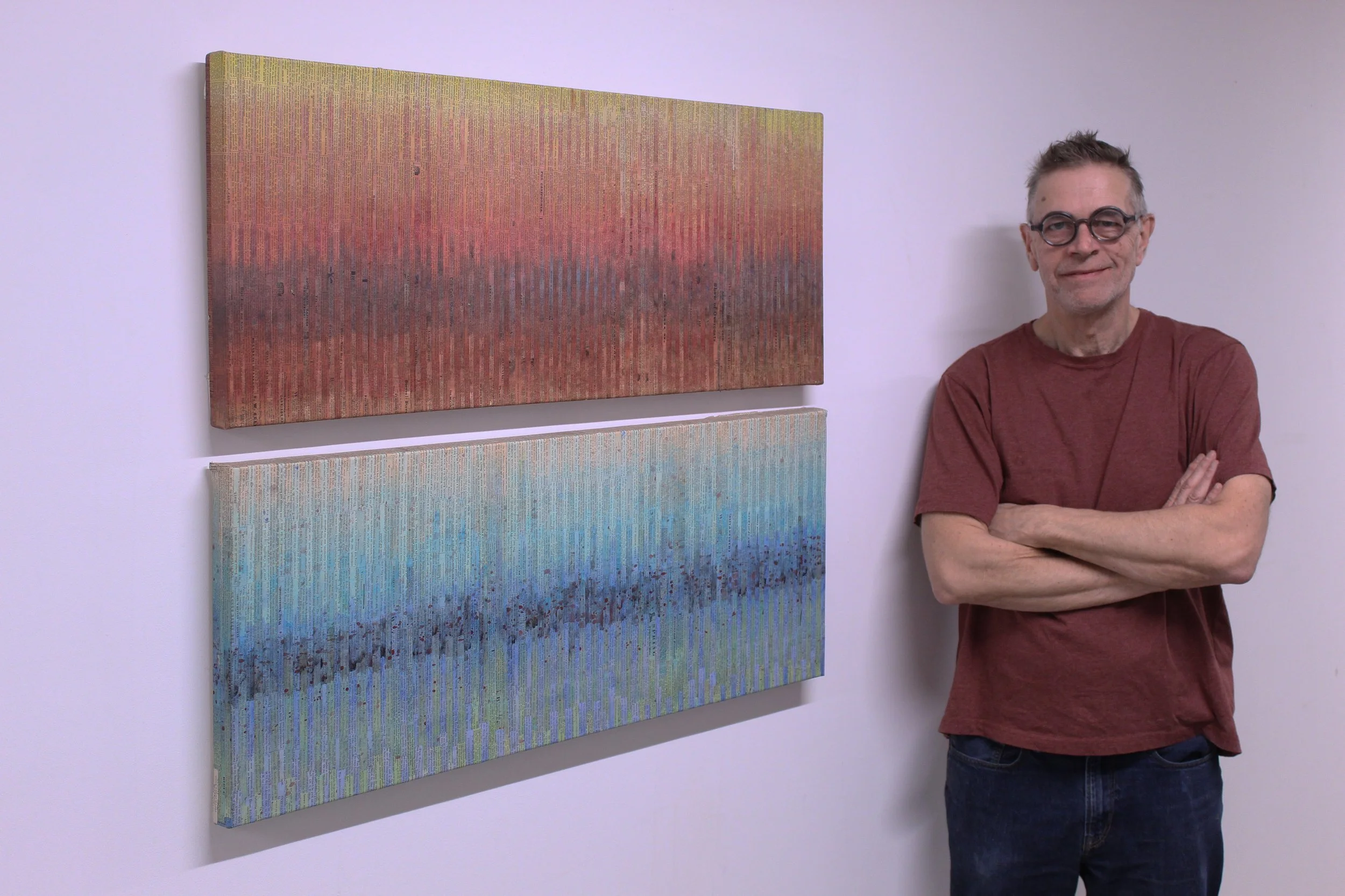 Chris Cloutier with glasses and a red t-shirt standing with arms crossed next to two abstract collage paintings on a white wall.