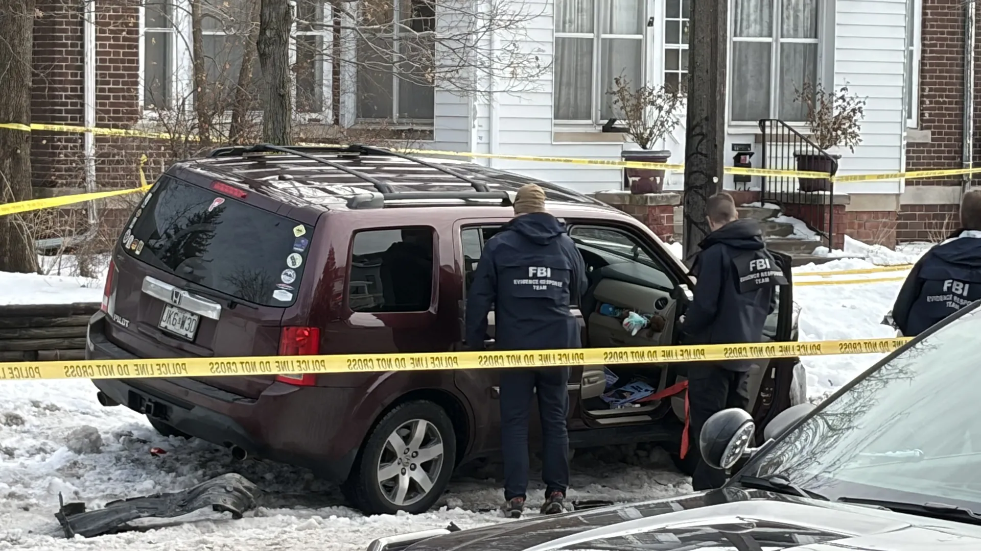 FBI agents inspecting a burgundy Honda Element vehicle behind yellow police tape in a residential neighborhood with snow on the ground and houses in the background.