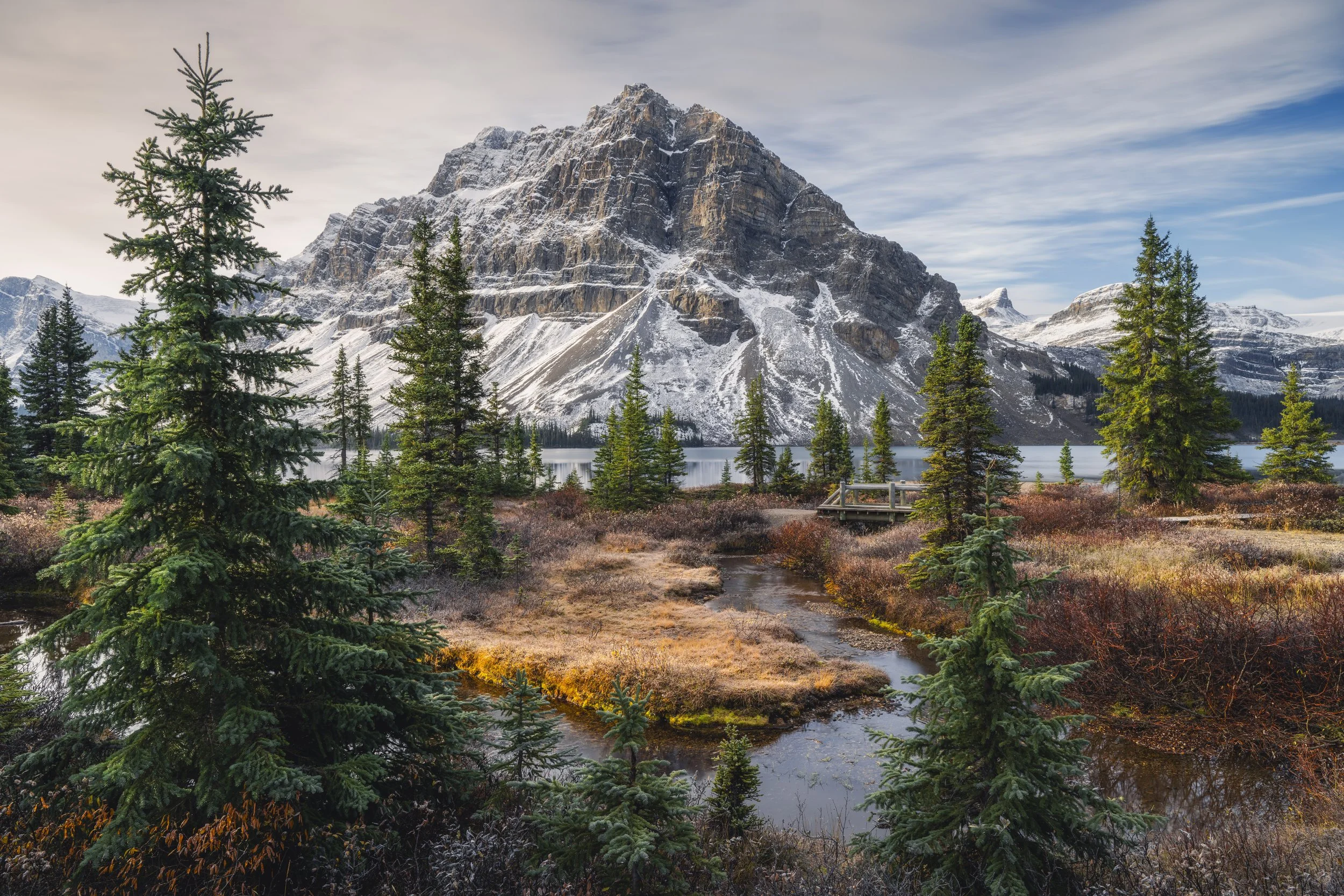 Snow-capped mountain behind evergreen trees near a lake with a small stream in the foreground, and a wooden observation deck on the lakeshore under a partly cloudy sky.