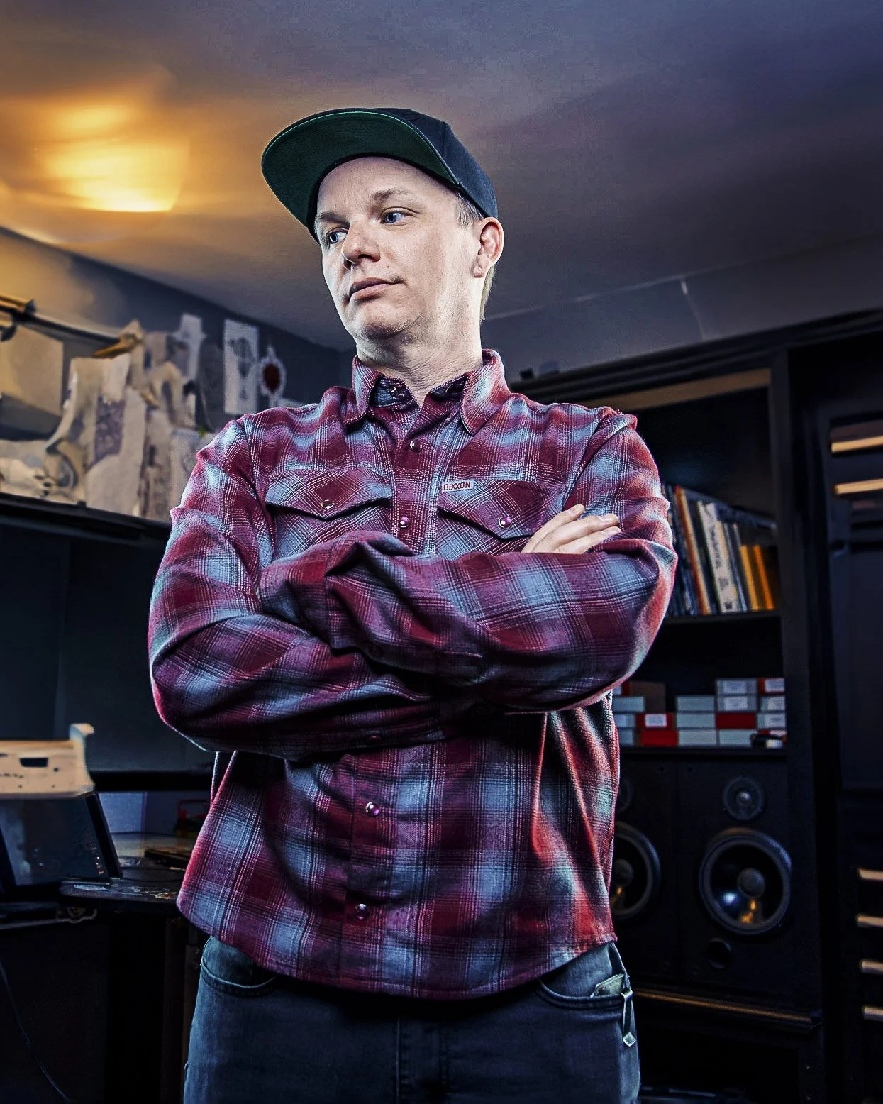 A young man with light skin, wearing a black baseball cap and a red plaid shirt, crossing his arms and looking to the side in a room filled with books and music equipment.
