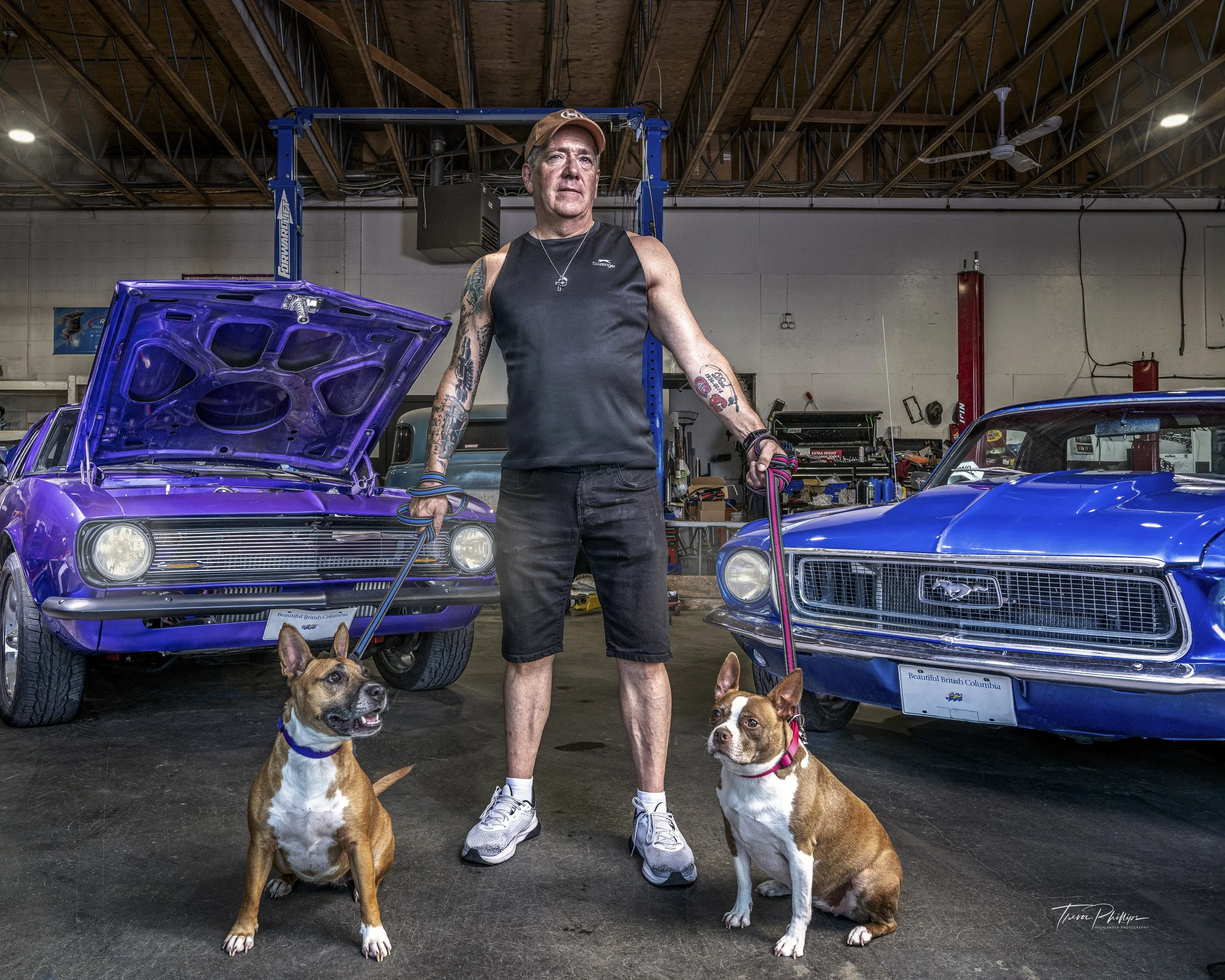 A man standing in an auto repair garage, holding two dogs on leashes, with two classic cars, one purple with its hood up and one blue, in the background.