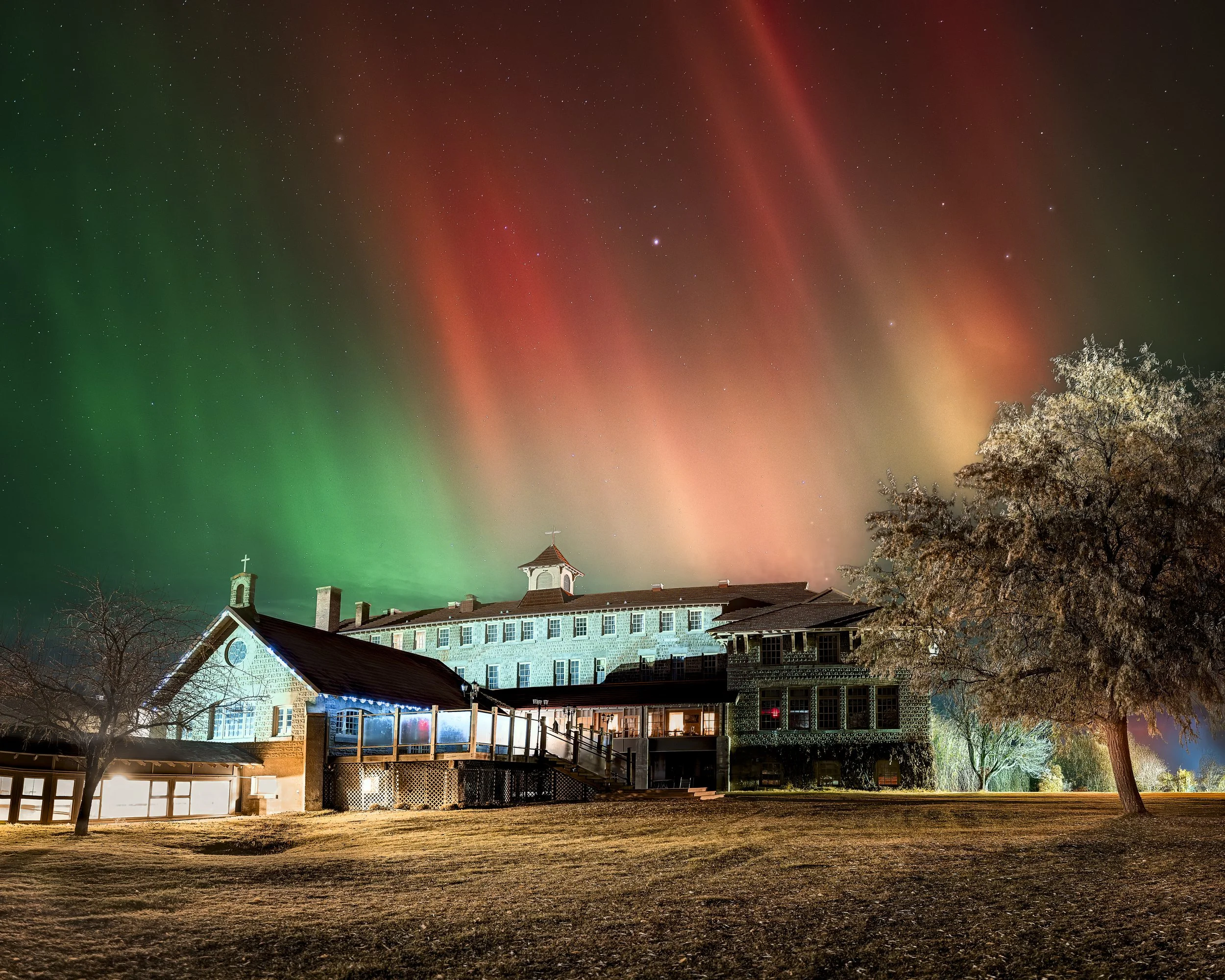 A large house with multiple stories and a steep roof, illuminated at night, under a colorful aurora borealis sky with green, red, and orange lights, and a starry sky in the background. There are trees in the foreground and a grassy area near the hous