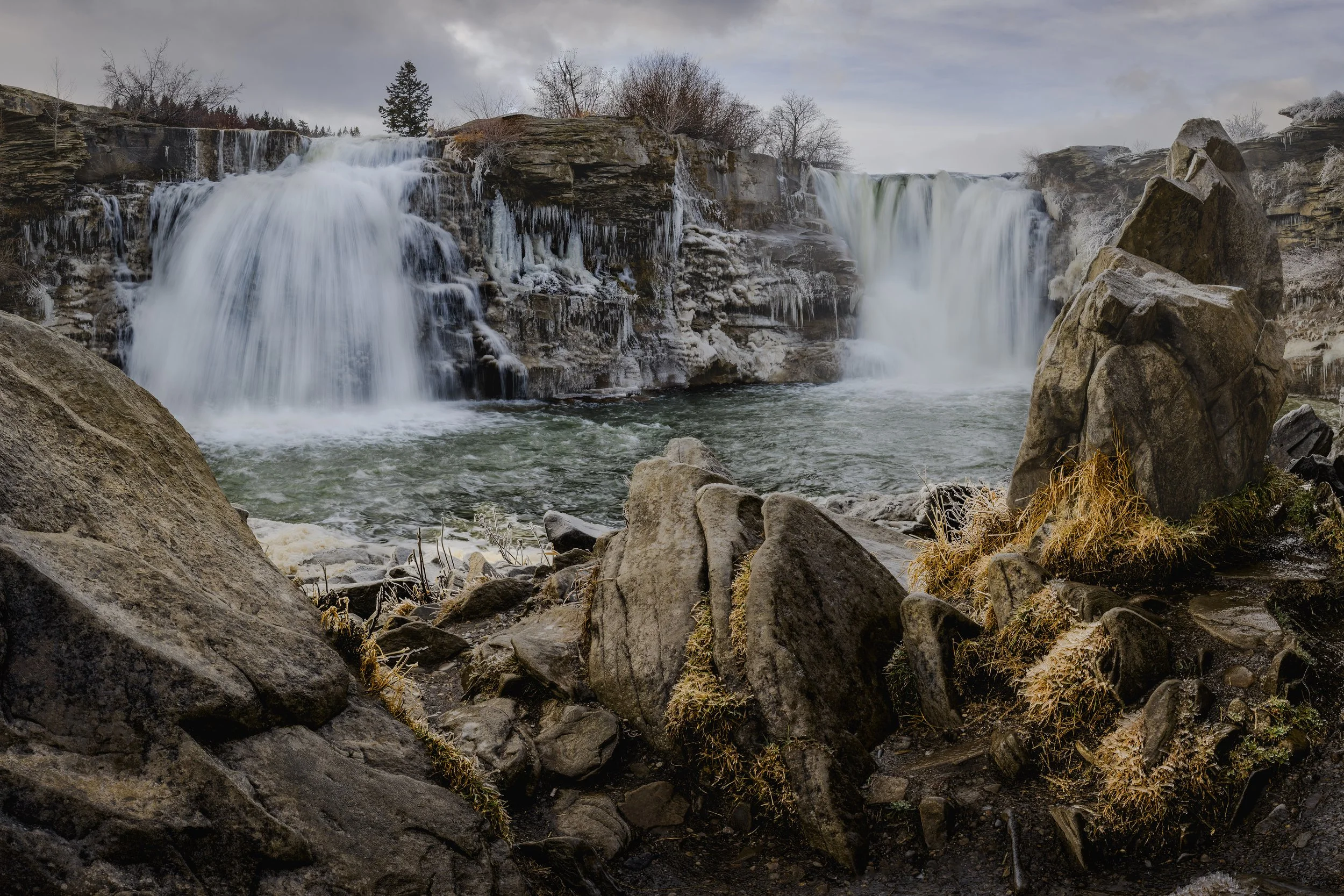 Waterfalls flowing over rocks in a natural landscape with leafless trees and cloudy sky.