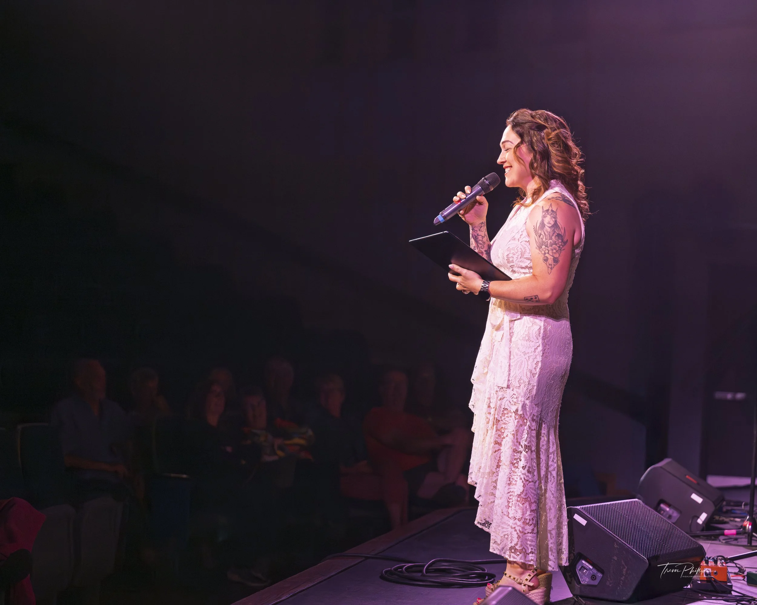 A woman in a pink lace dress standing on stage, holding a microphone and a tablet, performing or speaking to an audience in a dark room with stage lighting.