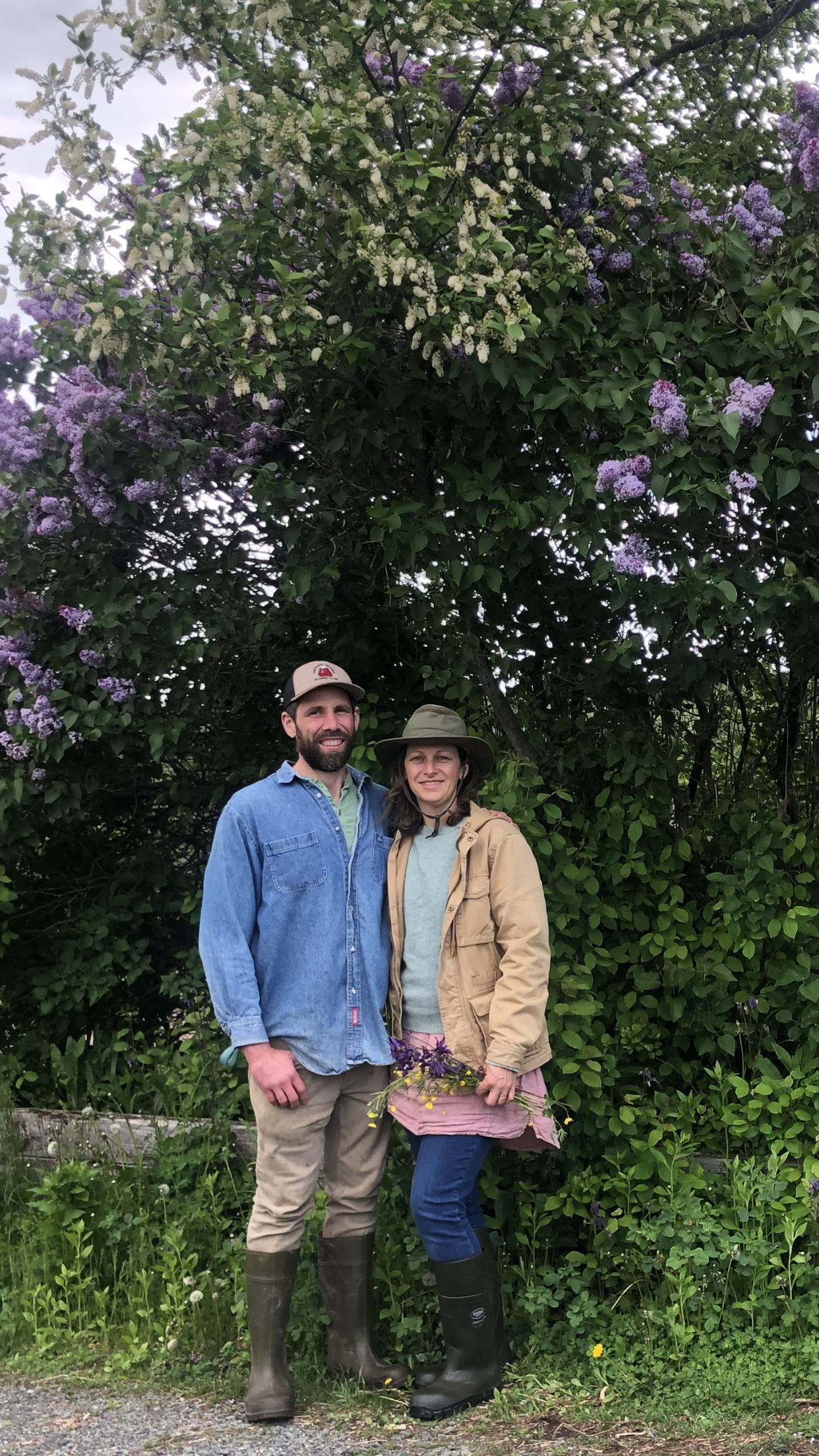 Suzanna and Justin standing outdoors in front of a large leafy bush with purple and white flowers at Ursa Farm,