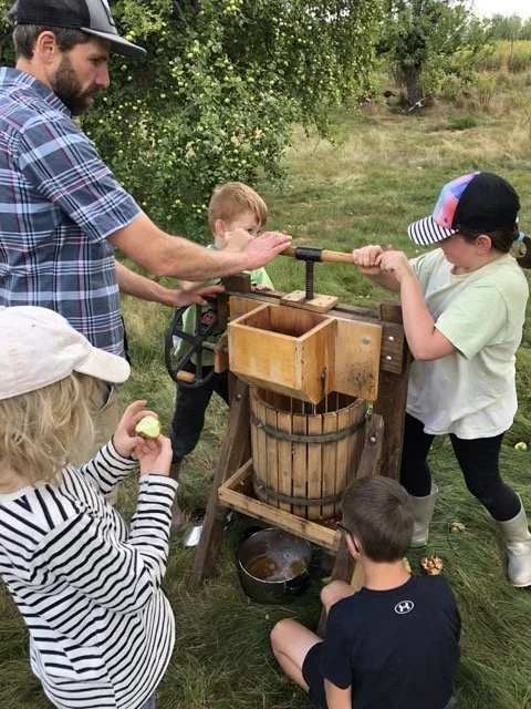 A group of children and an adult are outdoors, operating a vintage apple press to make cider. The children are turning the handle and holding apples, with trees and grass in the background.