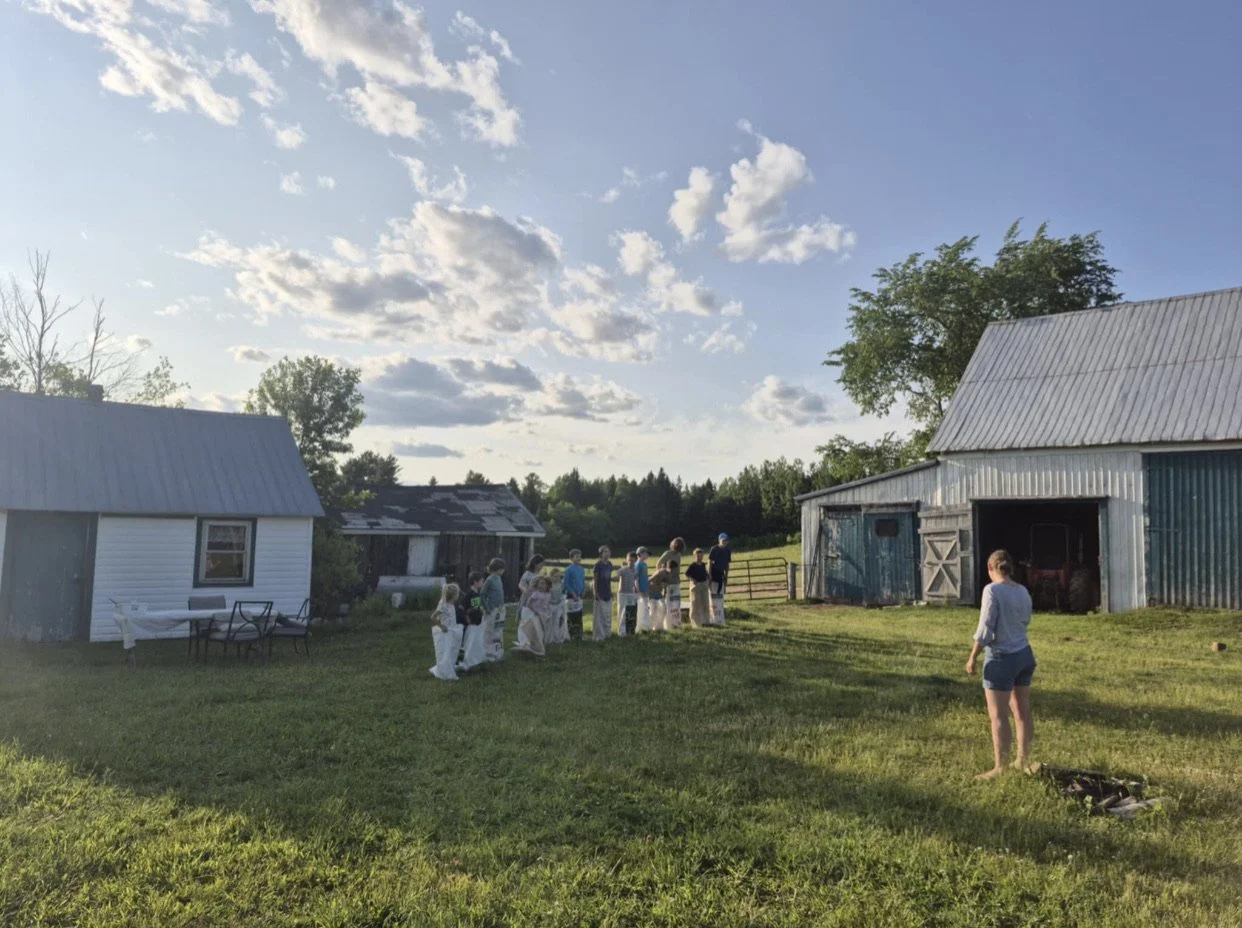 People participating in a sack race on a grassy farmyard with barns and trees, under a partly cloudy sky.