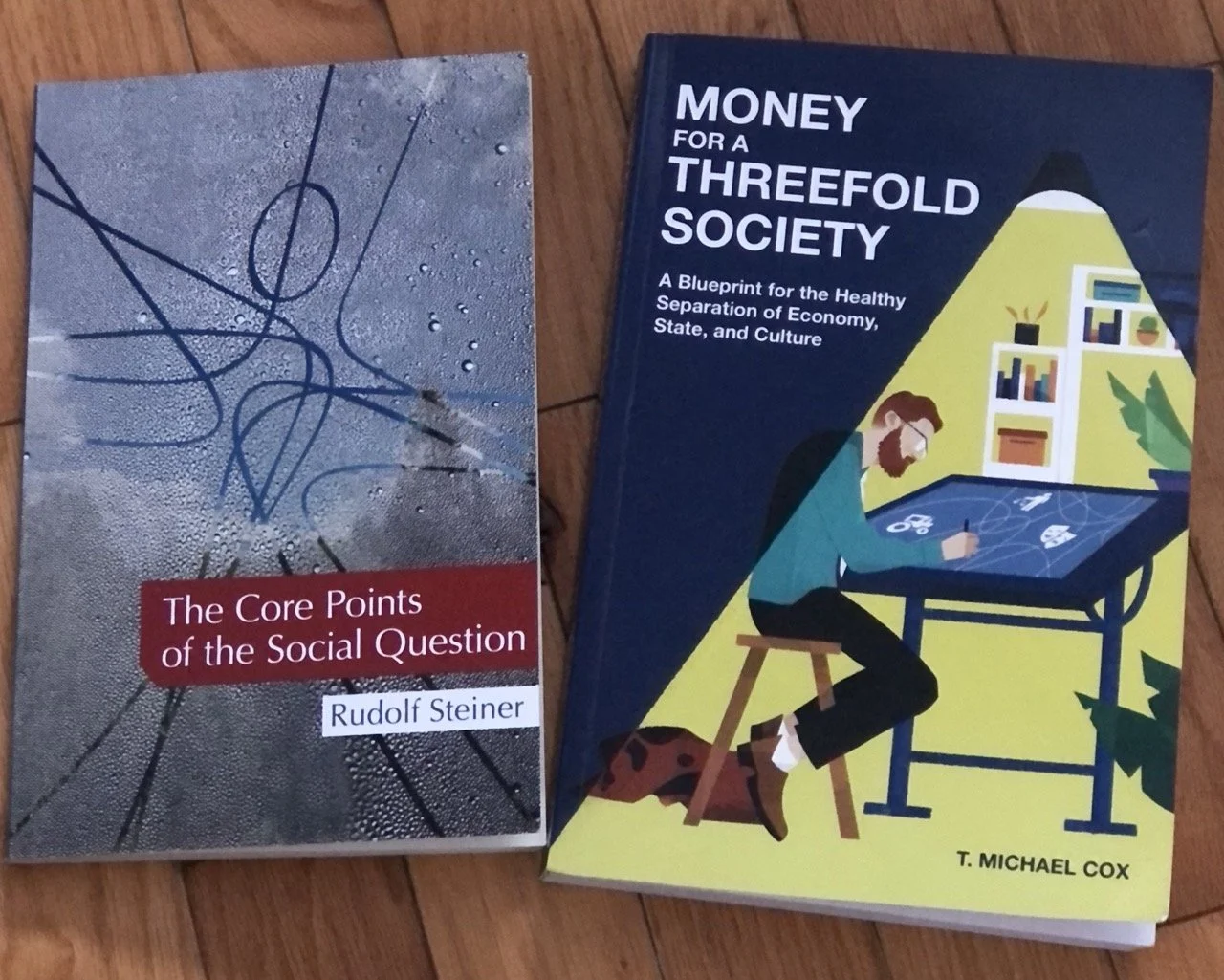Photo of two books placed on a wooden surface. The book on the left is titled "The Core Points of the Social Question" by Rudolf Steiner, with a textured gray cover featuring abstract black lines. The book on the right is titled "Money for a Threefold Society" by T. Michael Cox, with a dark blue and yellow cover illustration of a person using a tablet at a desk, illuminated by a lamp.