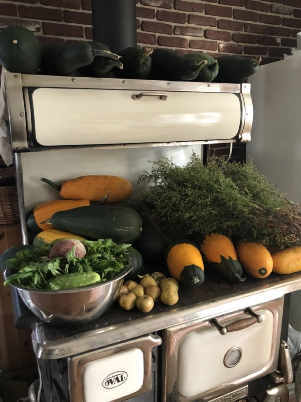 Fresh vegetables including zucchini, yellow squash, pumpkins, and herbs on a vintage stove with a brick wall background.