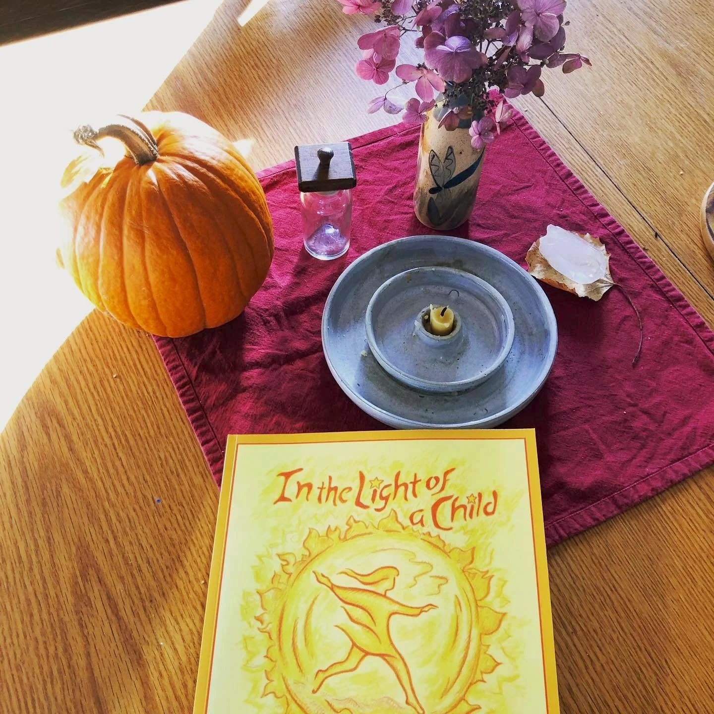 A Waldorf-themed table decorated for with a pumpkin, a red table runner, a vase with purple flowers, a small glass jar, a gray candle holder with a candle, a shell, a booklet titled 'In the Light of a Child,' and a few small decorative items.
