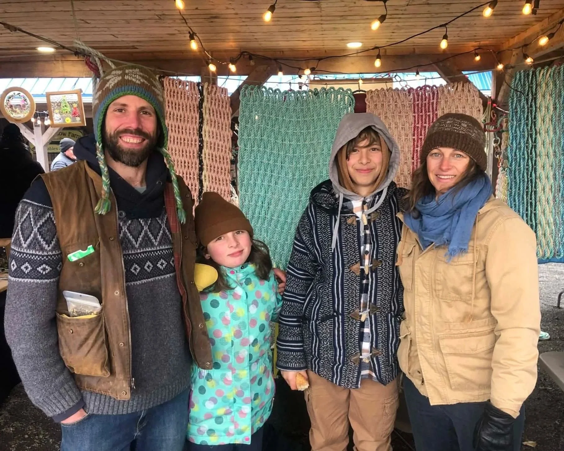 Ursa Farm family wearing warm clothing and hats, standing in front of colorful woven blankets, with string lights overhead.