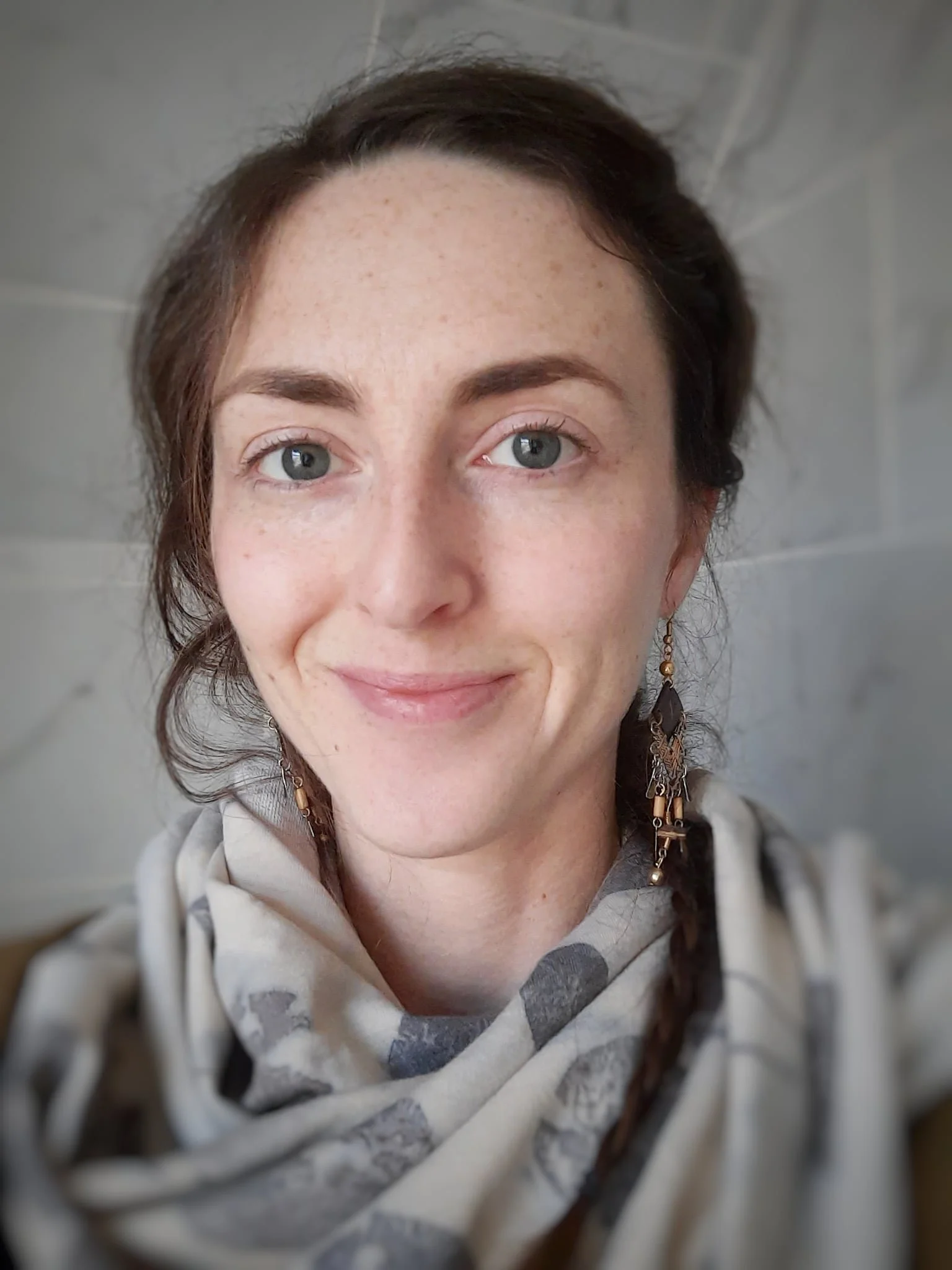 Close-up of a Layla with light skin, freckles, blue eyes, dark hair, wearing earrings and a patterned scarf, smiling in front of a gray tiled background.