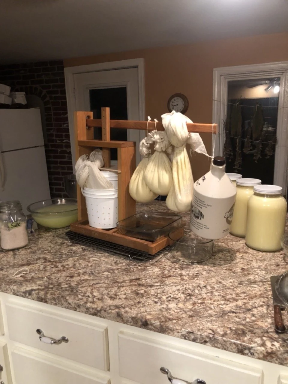 A kitchen countertop holding cheese and garlic bulbs; various jars and containers of dairy products, a bowl, a glass baking dish, and some kitchen utensils and supplies.