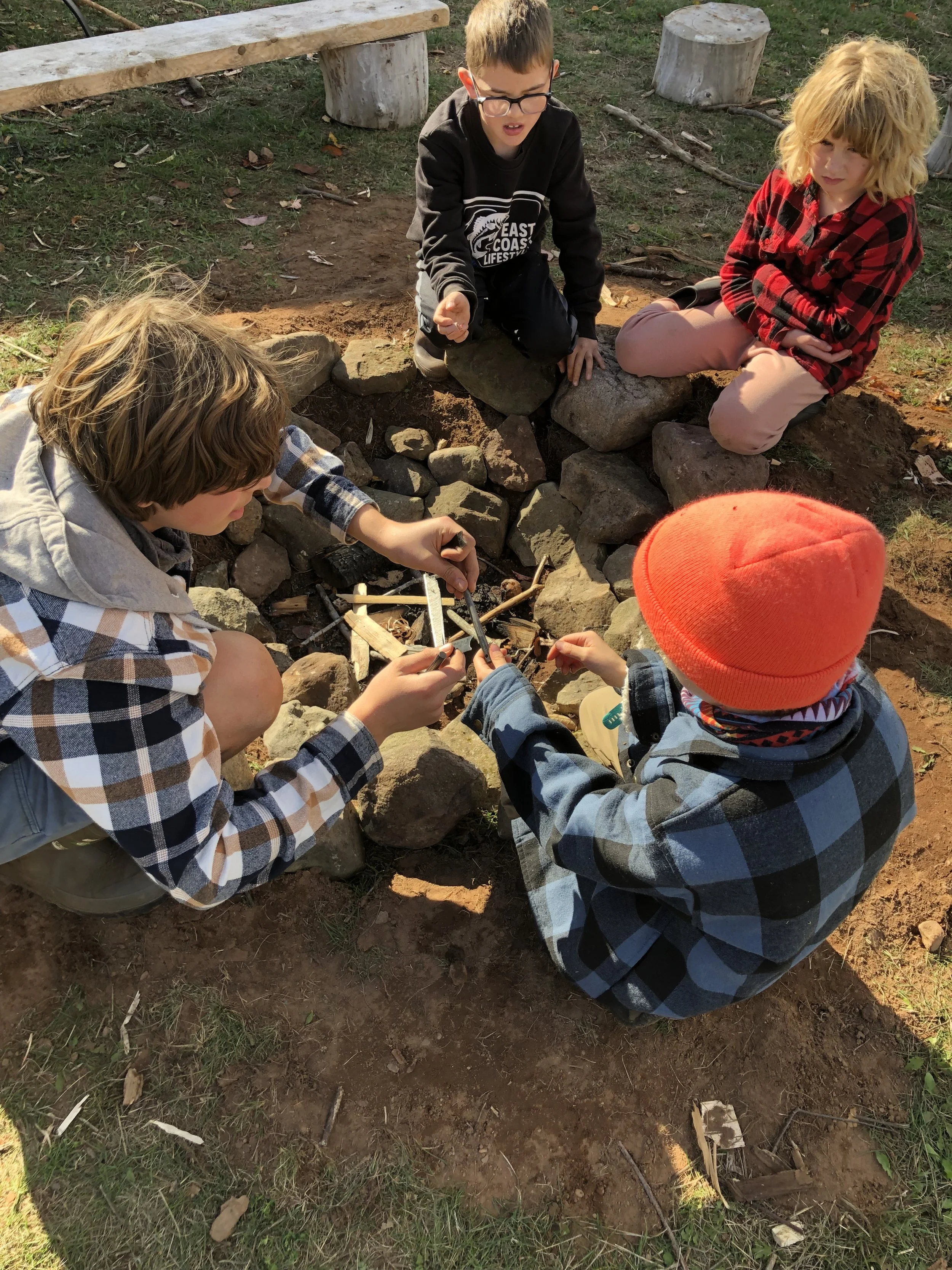 Children gathered around a small fire pit made of rocks, outside on dirt and grass, building or tending to a small campfire with sticks.