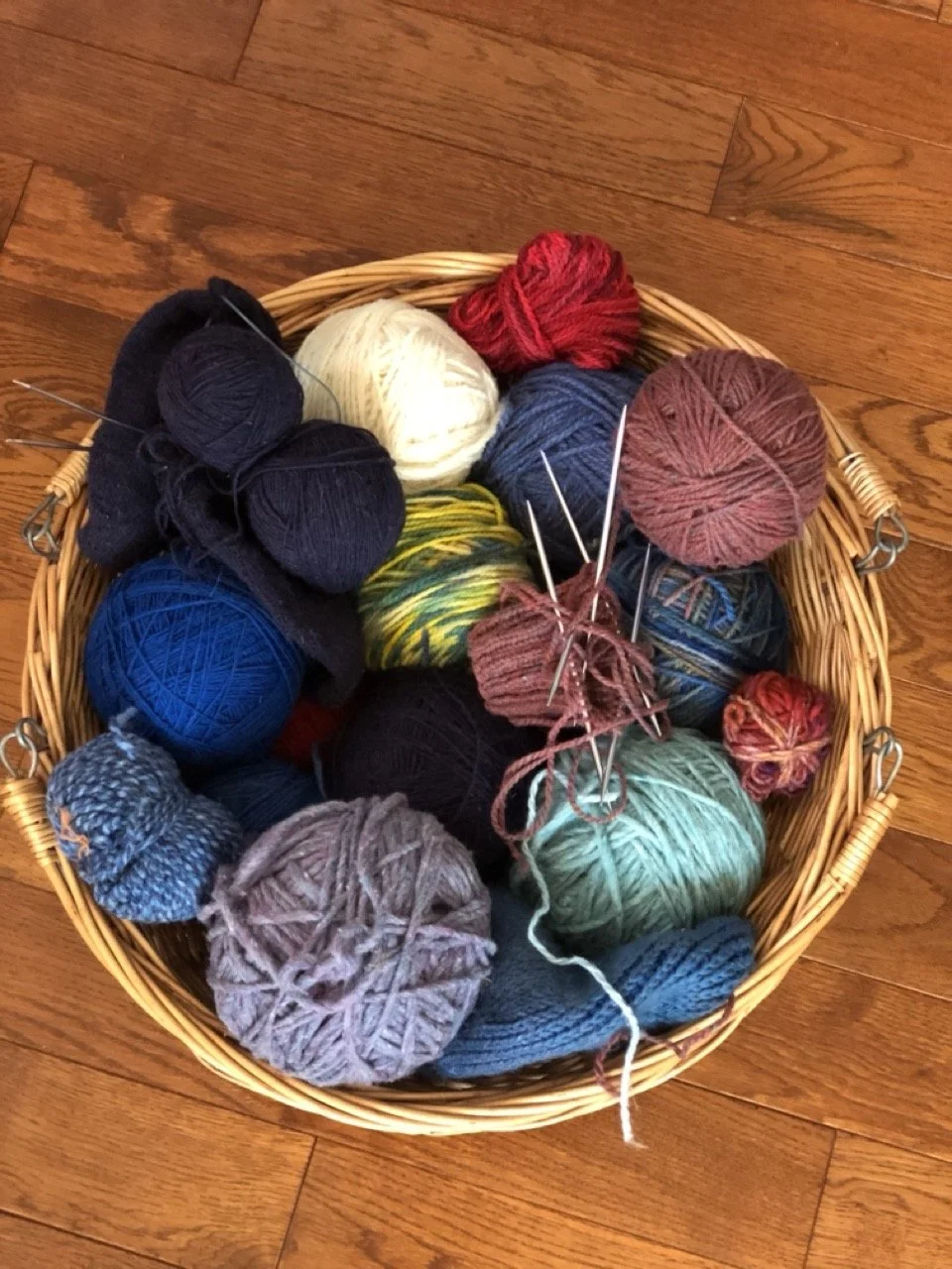 Basket filled with various colored balls of yarn, some with knitting needles, on a wooden floor.