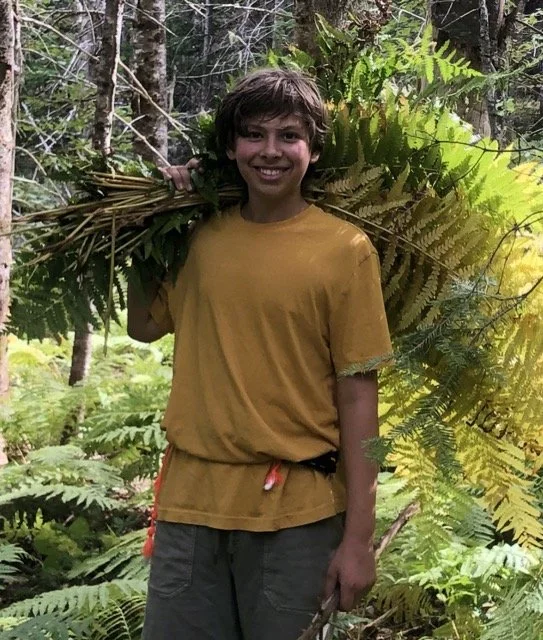 A smiling boy, Gabe, with curly hair wearing a yellow t-shirt and grey pants carrying a large bundle of green leaves on his shoulder in a lush forest.