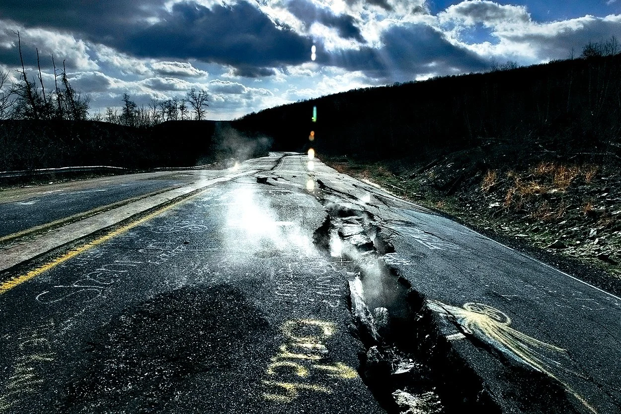 A damaged wet asphalt road with cracks and a large gouge along the center, reflected in the water puddles, in a hilly area under a cloudy sky with sunlight breaking through.