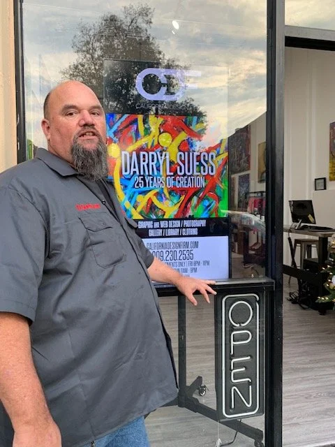 Man standing outside of an art gallery or studio with a colorful art poster behind him. The poster features the name Darryl Suess and mentions 25 years of creation. The man is wearing a gray shirt with red writing and has a beard. There is a black stand with a white logo or sign reading 'CALUZ' in front of him.
