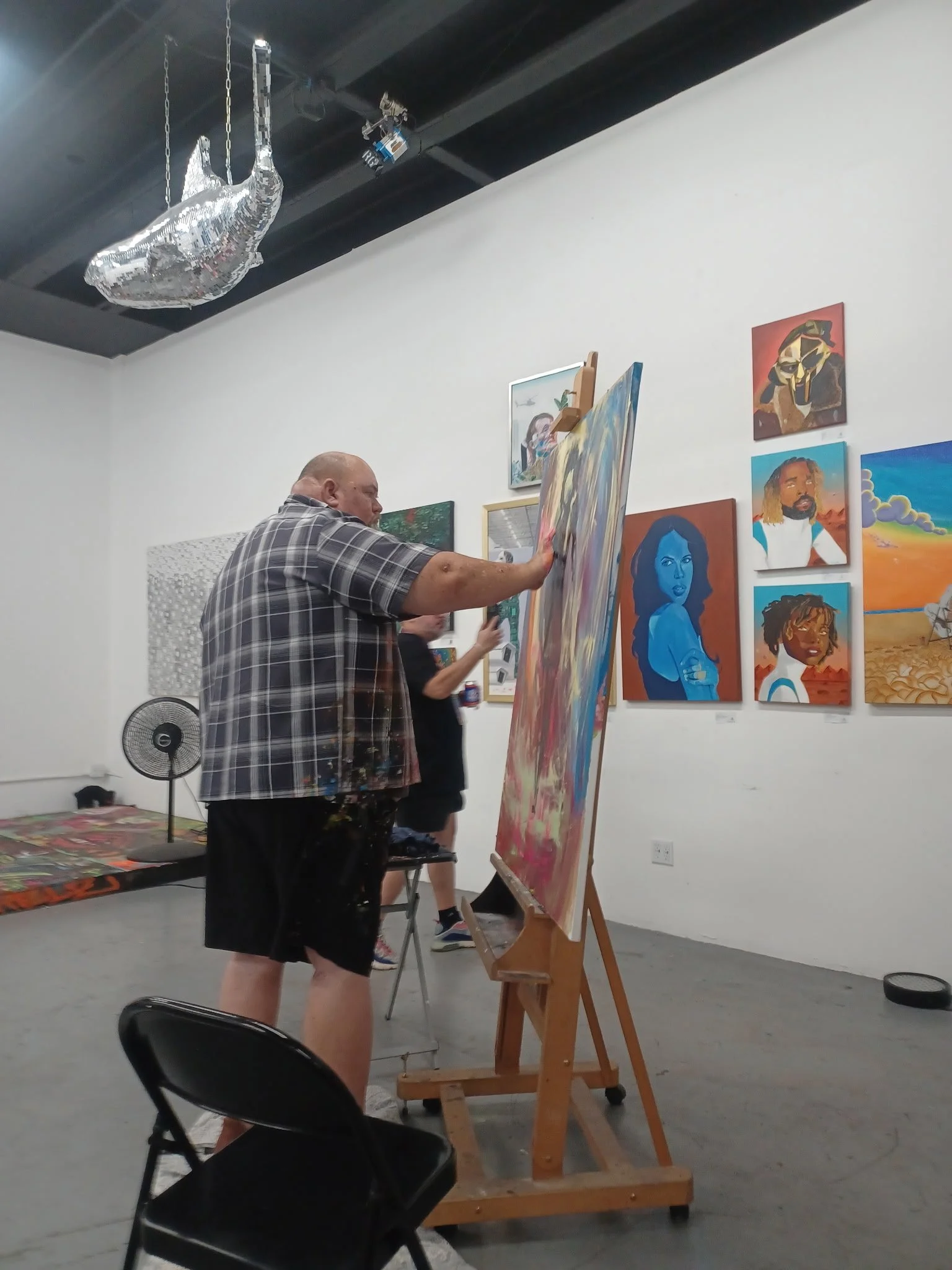 An artist in a plaid shirt is painting on an easel in an art gallery. The wall behind them displays various colorful portraits and artworks. A disco ball hangs from the ceiling.