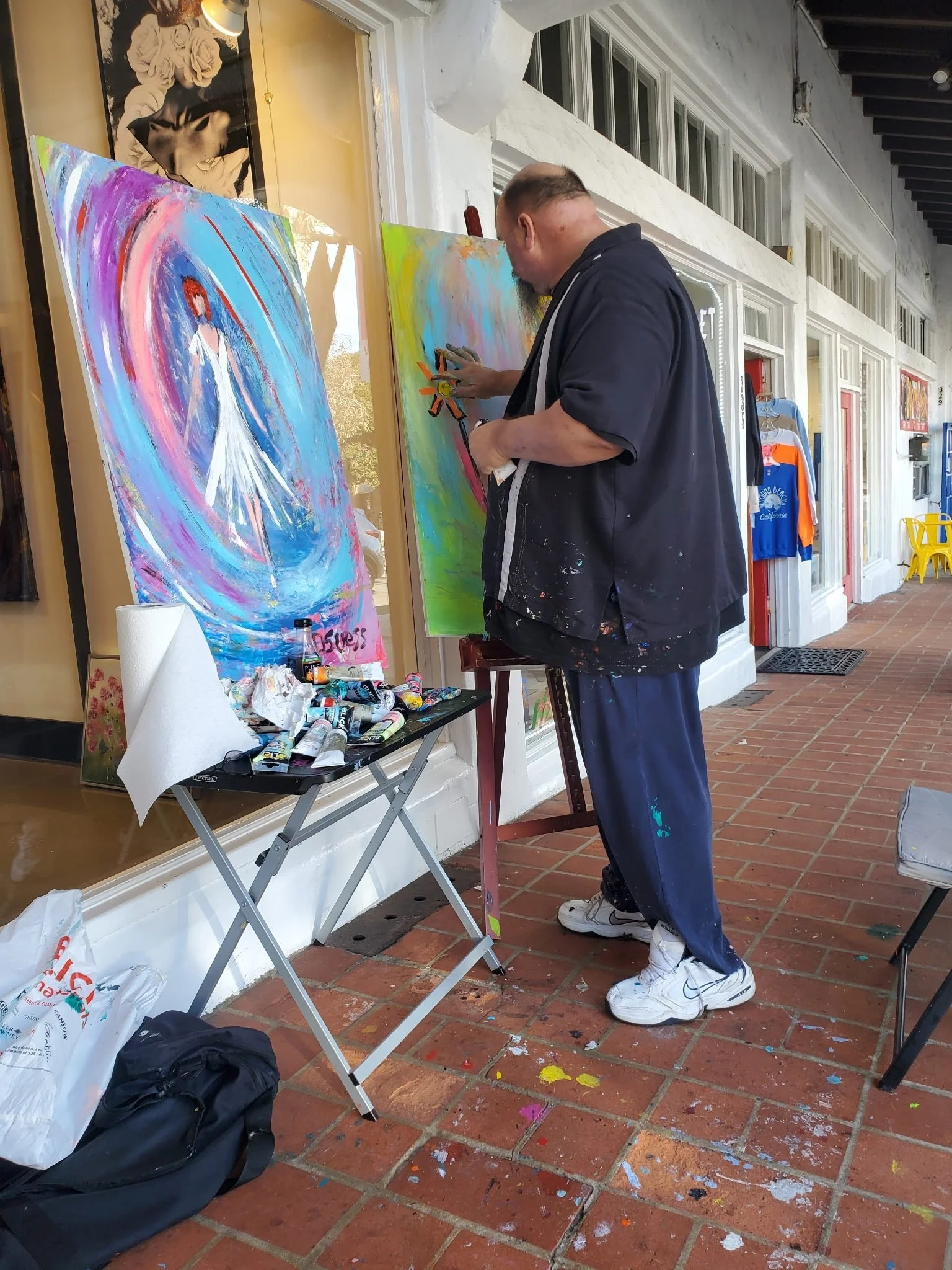 An artist painting outdoors on a sidewalk, with art supplies and colorful paintings visible, in front of a storefront with large windows and clothing displays.
