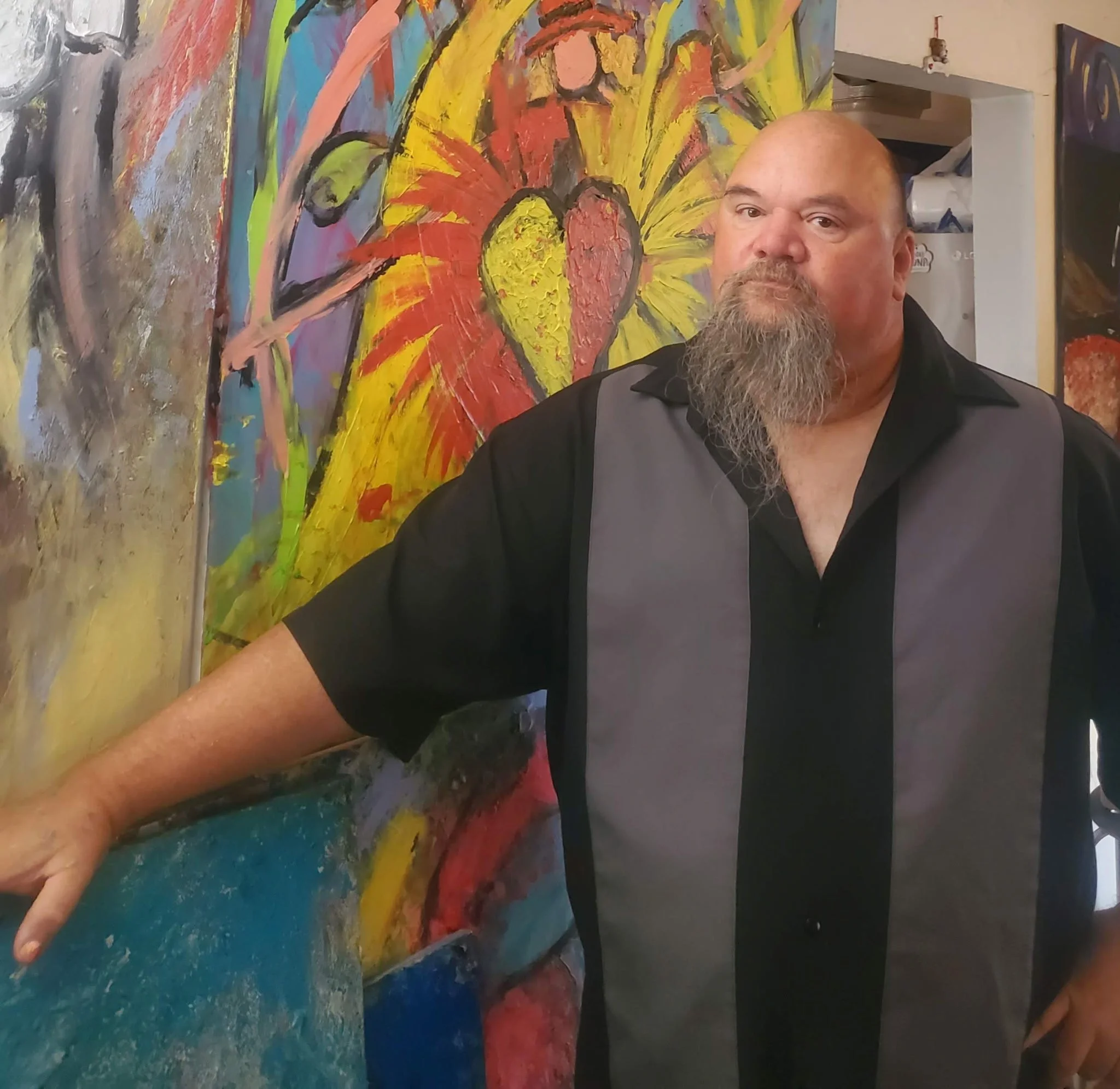 A person with a beard and wearing a black shirt is standing next to an abstract painting with vibrant yellow, red, purple, blue, and black colors. The person is in an indoor setting.