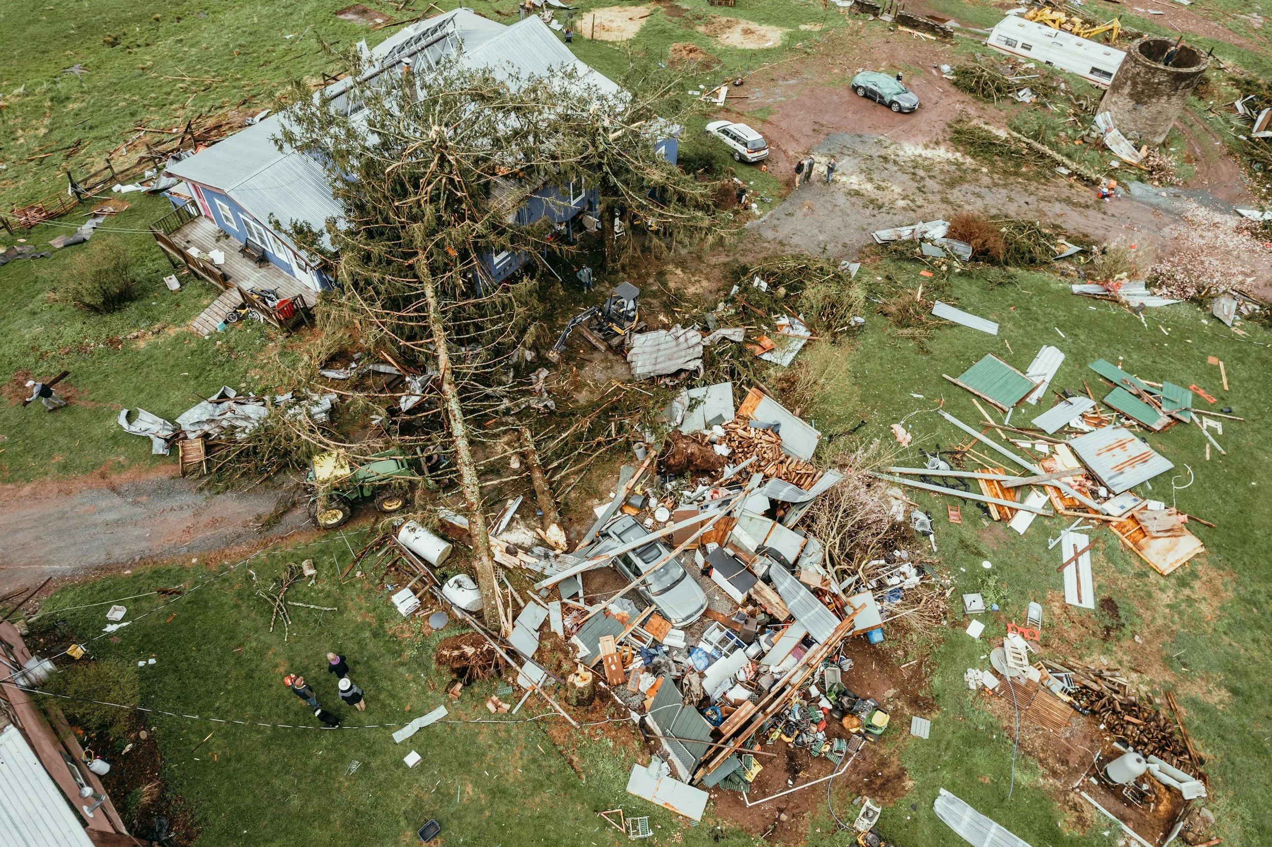 Aerial view of a damaged house with fallen trees, debris, and scattered roofing materials after a storm or tornado.