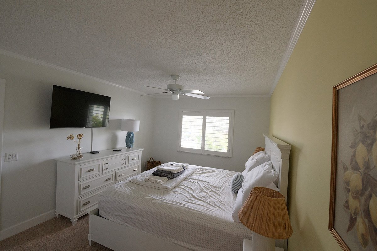 A neatly made bedroom with a white bed, a wicker bedside lamp, a white dresser with decorative items, and a window with blinds.