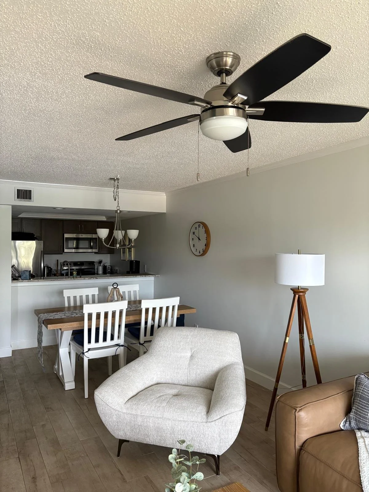 Living room with ceiling fan, white armchair, wooden floor, floor lamp, and dining area with white chairs and a wooden table, kitchen in background.