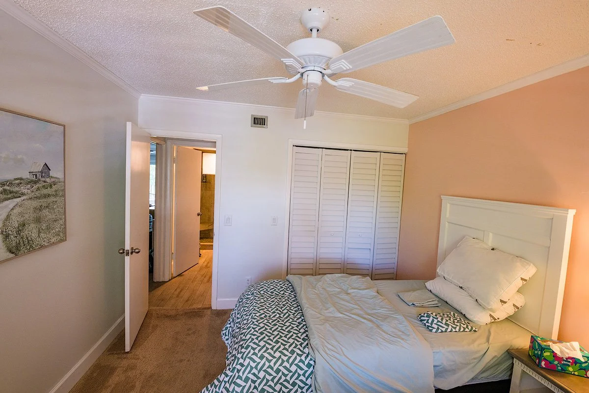 A bedroom with beige and white walls, a white bed with a large pillow, a patterned blanket, a small nightstand with a colorful box, a white ceiling fan, a closet with louvered doors, and an open door to a bathroom.
